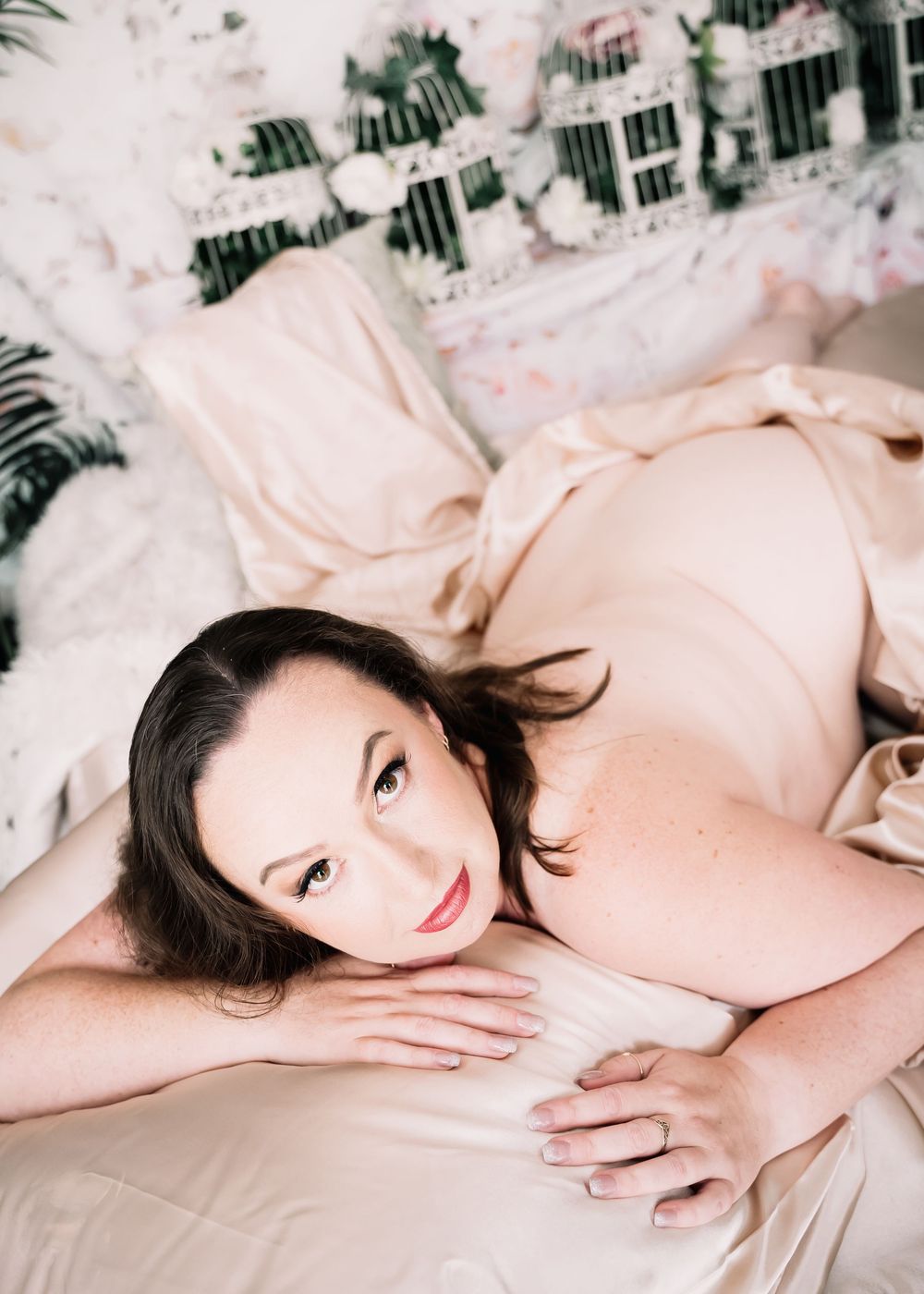 Woman in soft pink maternity attire reclines on light bedding while elegant pregnancy photos are taken.
