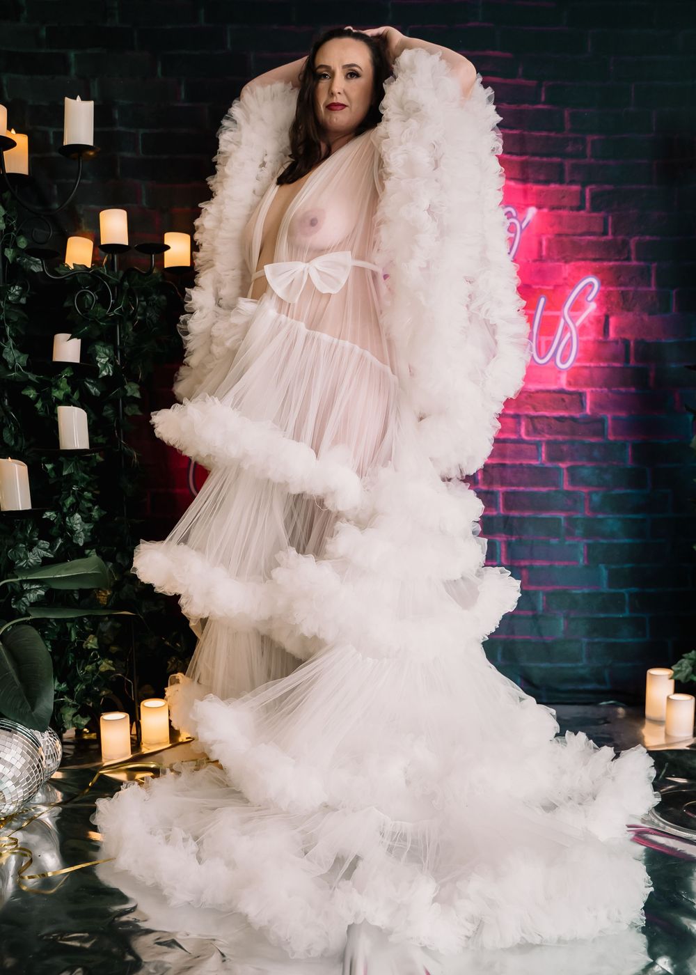 A dreamy boudoir photoshoot with ethereal white tulle fabric and glowing candles against a dark brick wall.