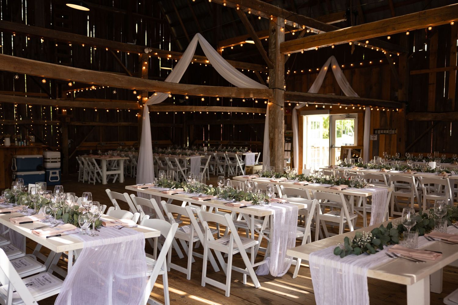 Long wooden table with white linens and rustic decor inside a barn wedding reception venue with twinkle lights overhead.