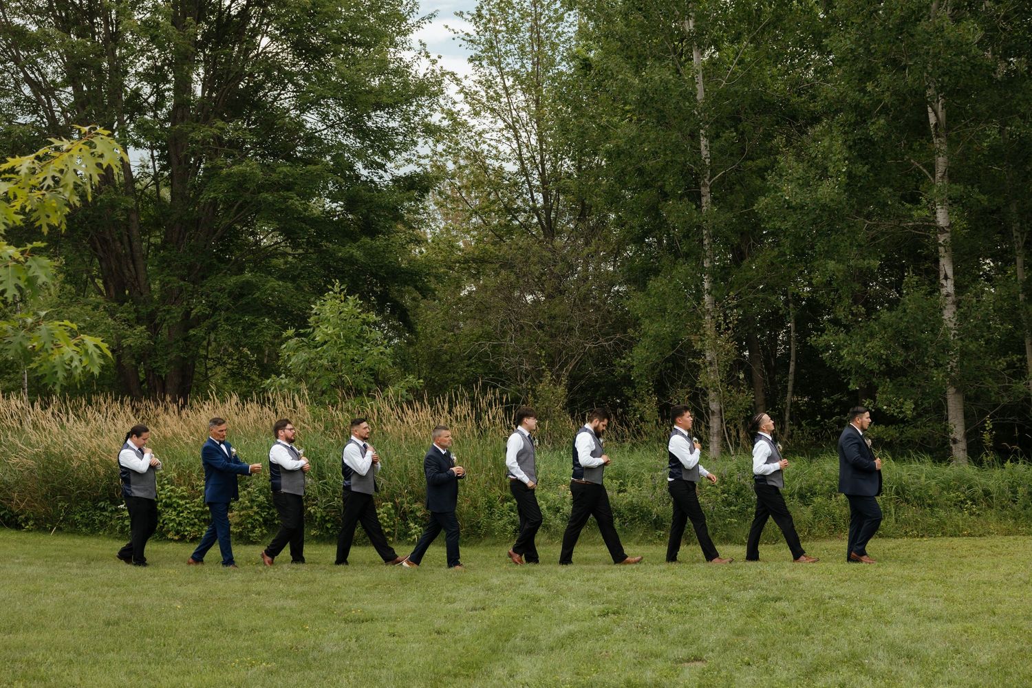 Wedding party in formal attire walks in line formation across a lush green lawn surrounded by trees on a sunny day.