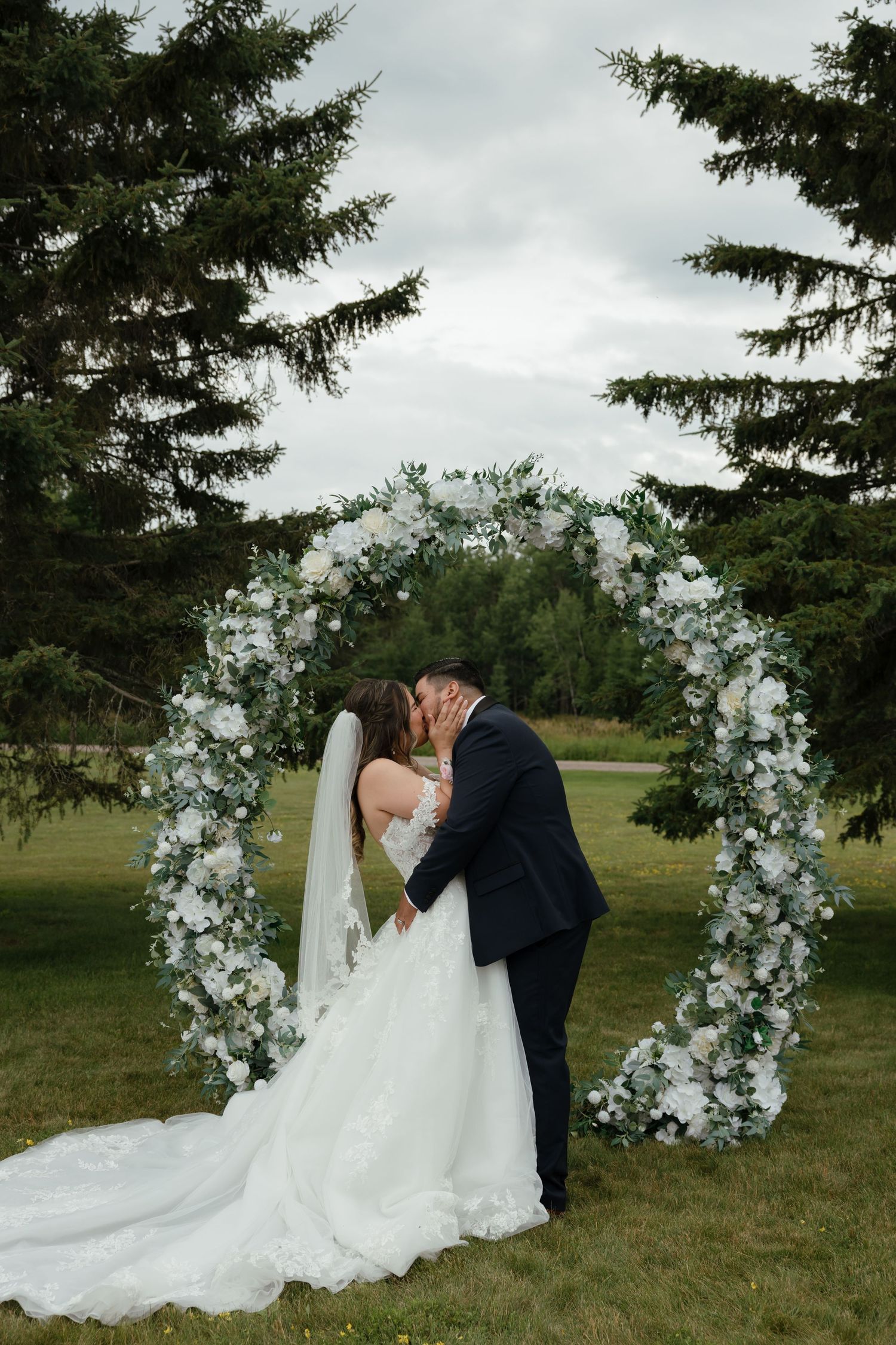 Romantic wedding couple shares tender moments under floral arch against natural green outdoor setting.