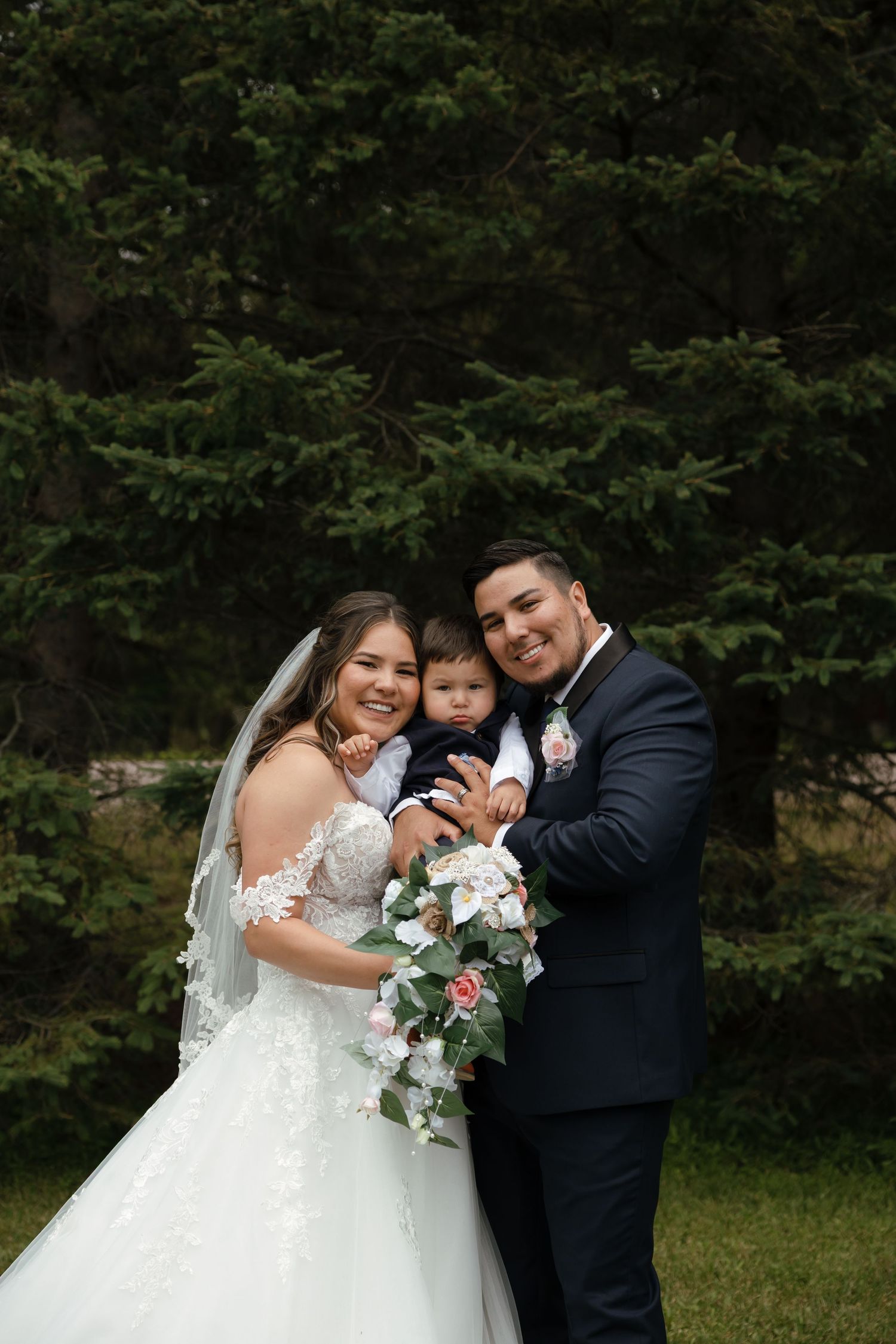 A joyful outdoor wedding photoshoot shows a family laughing together in a garden setting with white florals and formal attire.