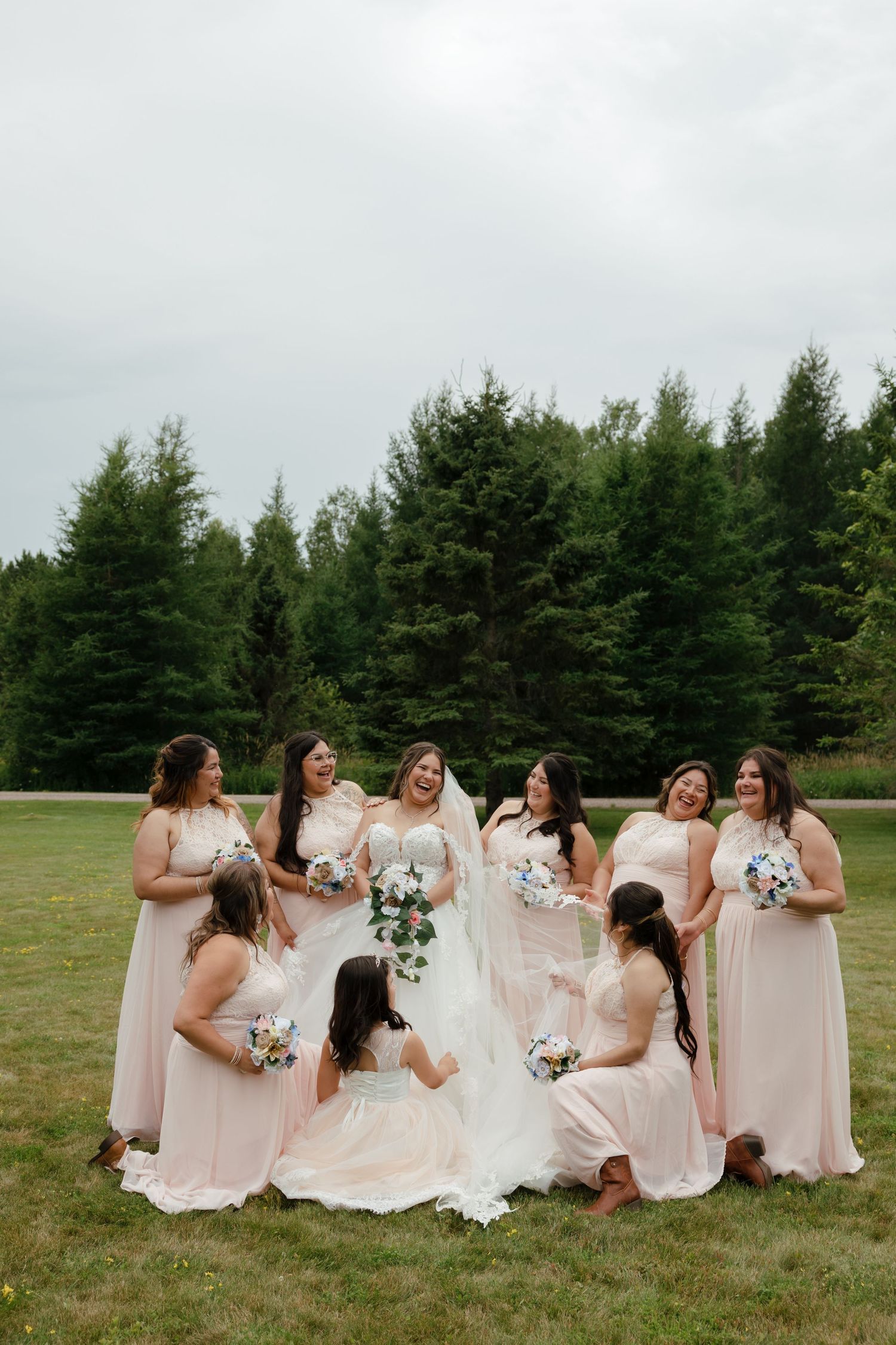 Wedding party in blush pink dresses sits casually on grassy field with bouquets sharing candid laughter together.