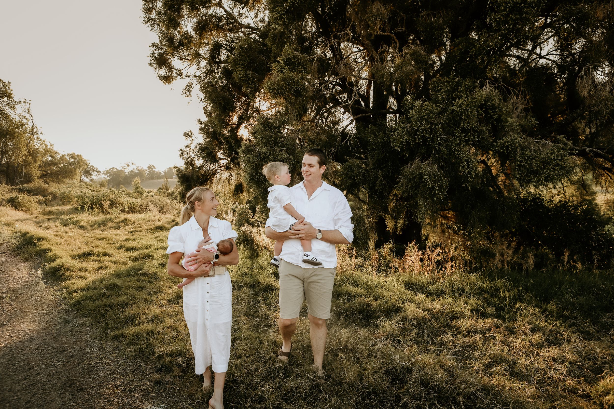 A couple in white outfits poses together during a golden hour outdoor photoshoot in a rustic natural setting.