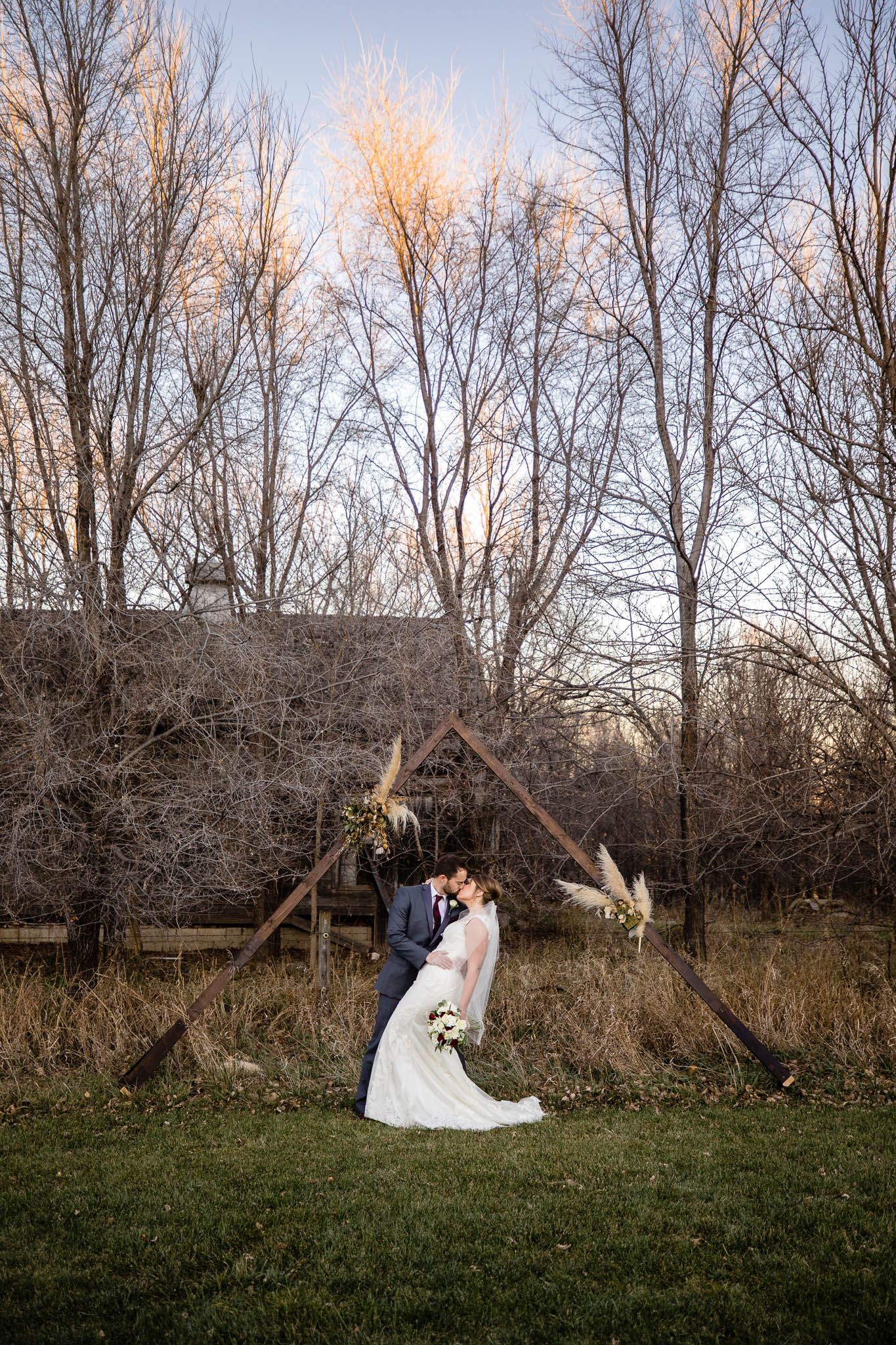 A couple embraces against a rustic wooden country fence at sunset with bare trees in the background.