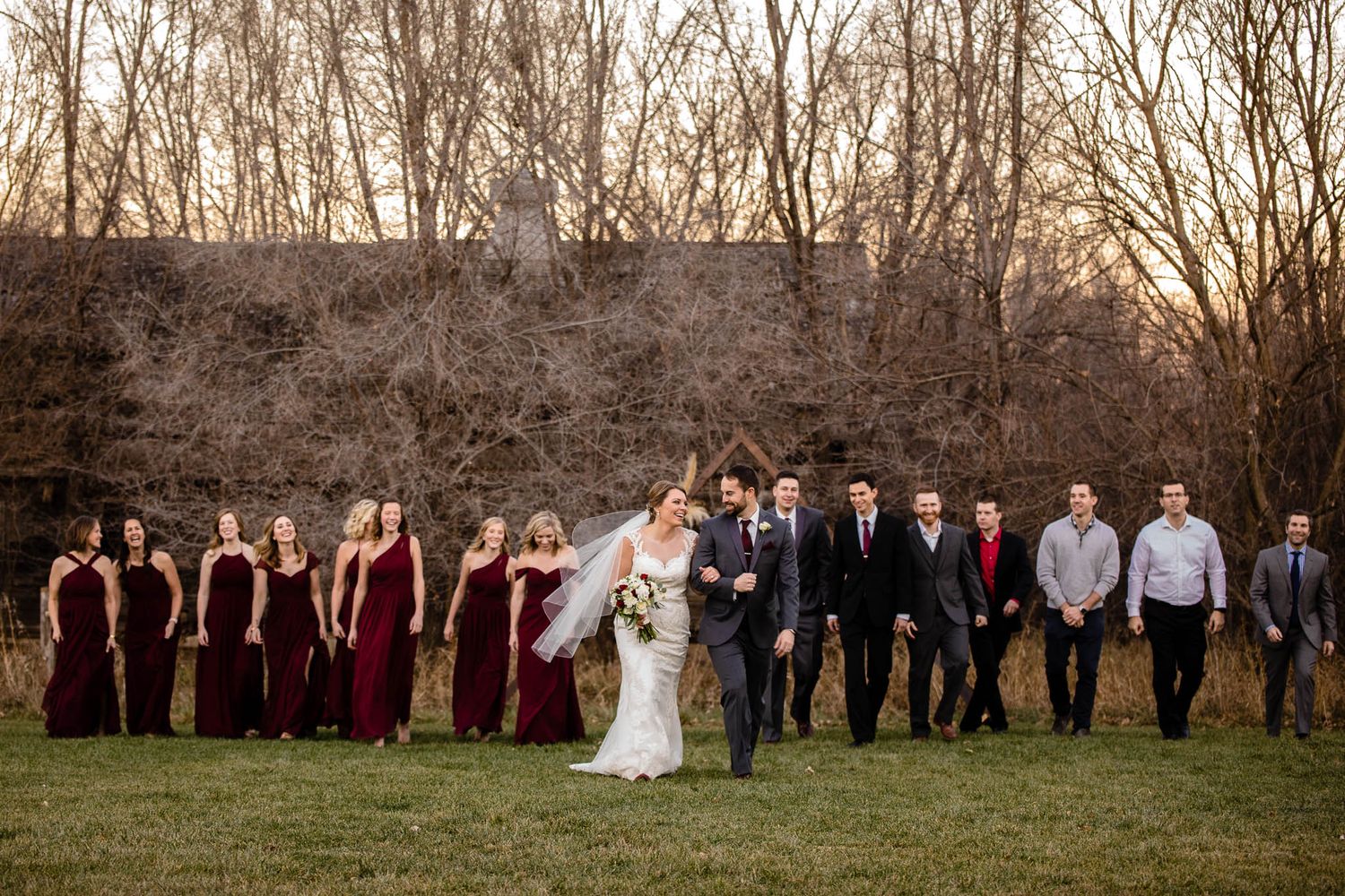 Elegant wedding party in maroon and black attire poses together outdoors at sunset during fall.