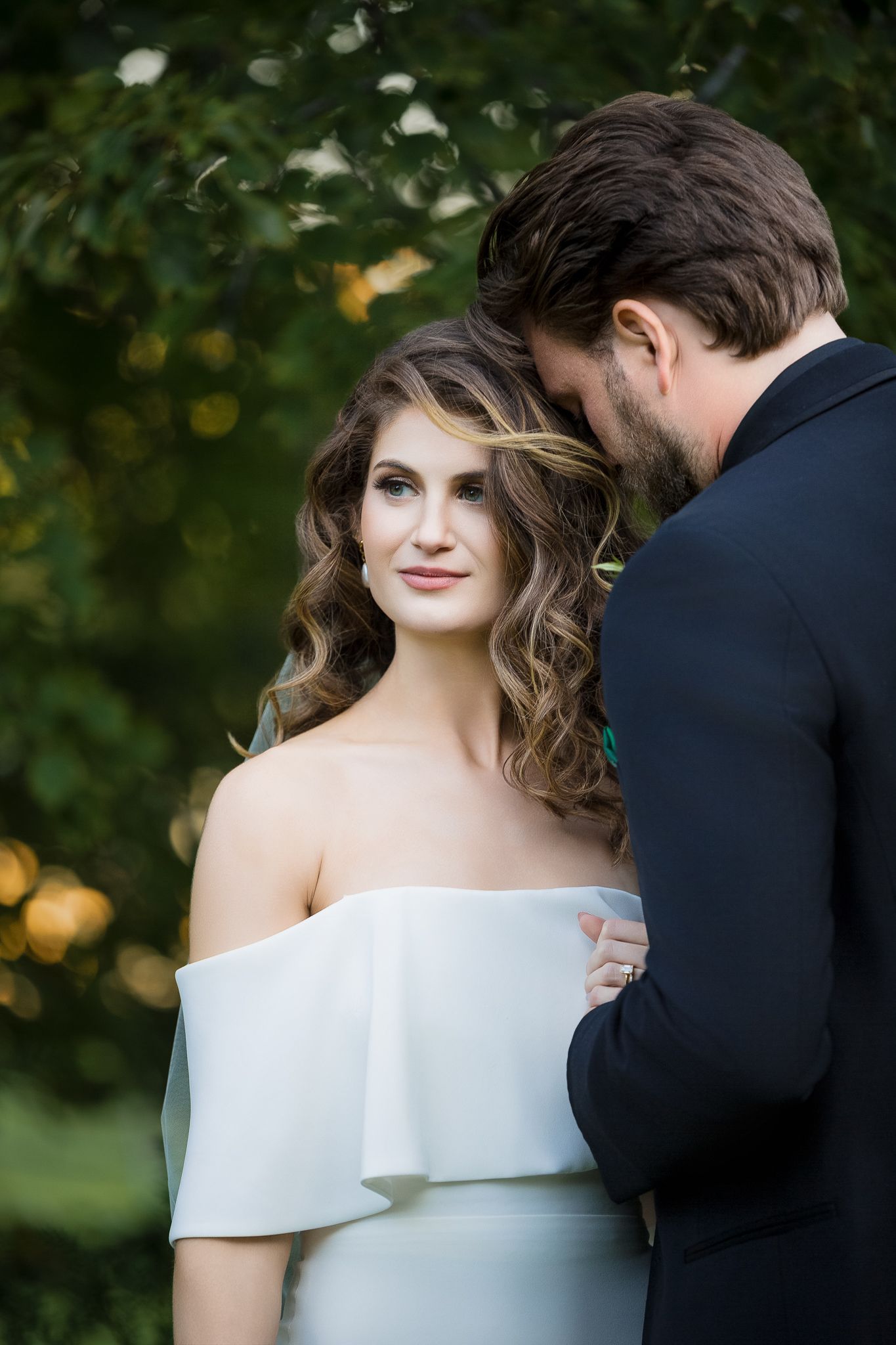 A romantic wedding couple embraces outdoors with soft natural lighting and greenery in the background.