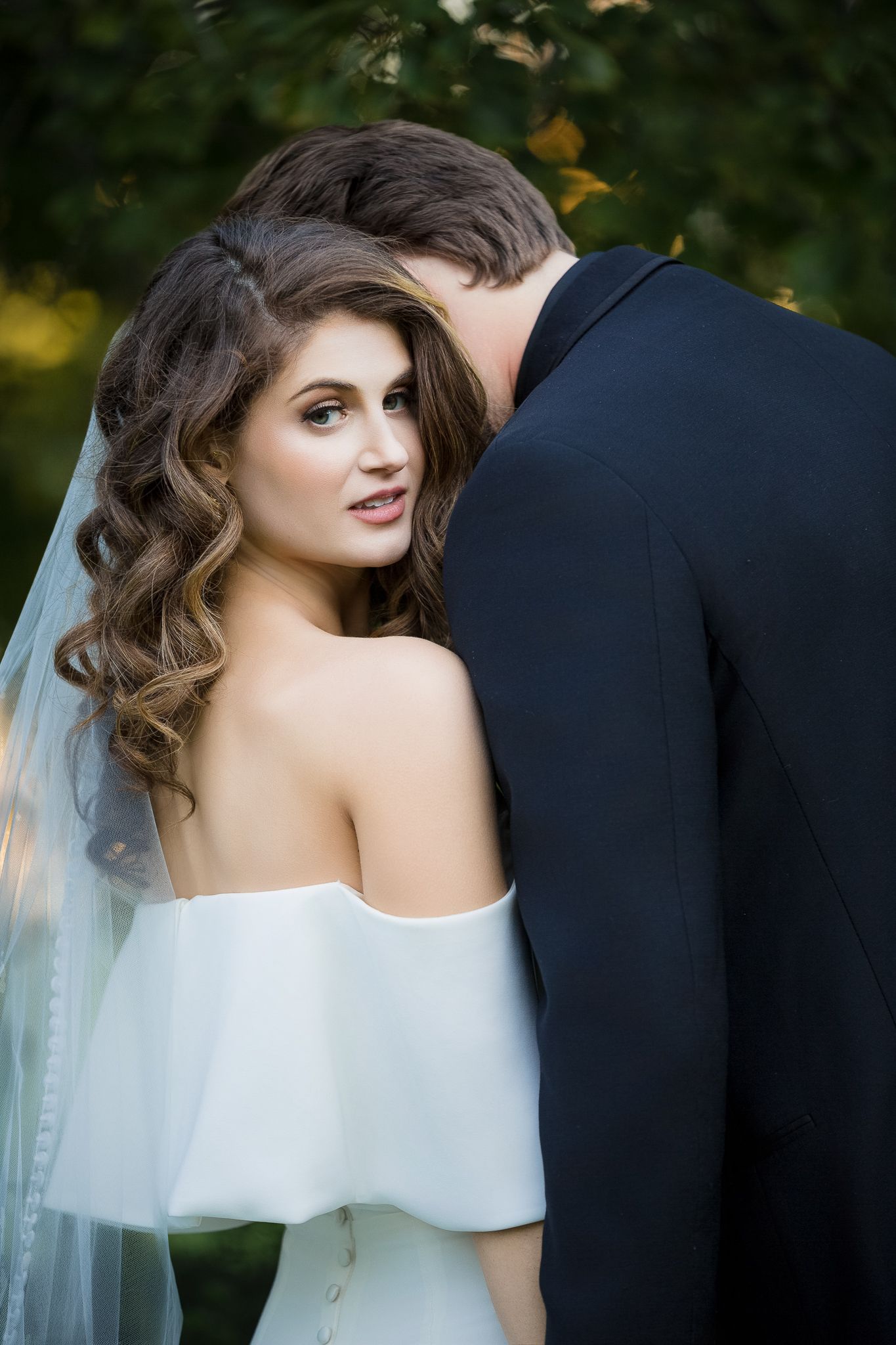 Romantic wedding portrait showing bride in off-shoulder white dress with flowing veil and dark curled hair outdoors.