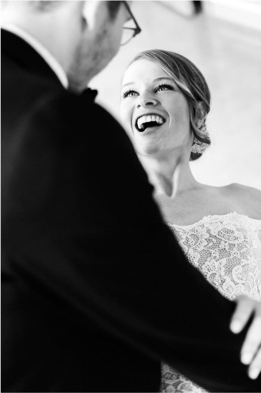 Black and white wedding photo captures intimate moment between bride in lace gown and groom during ceremony.