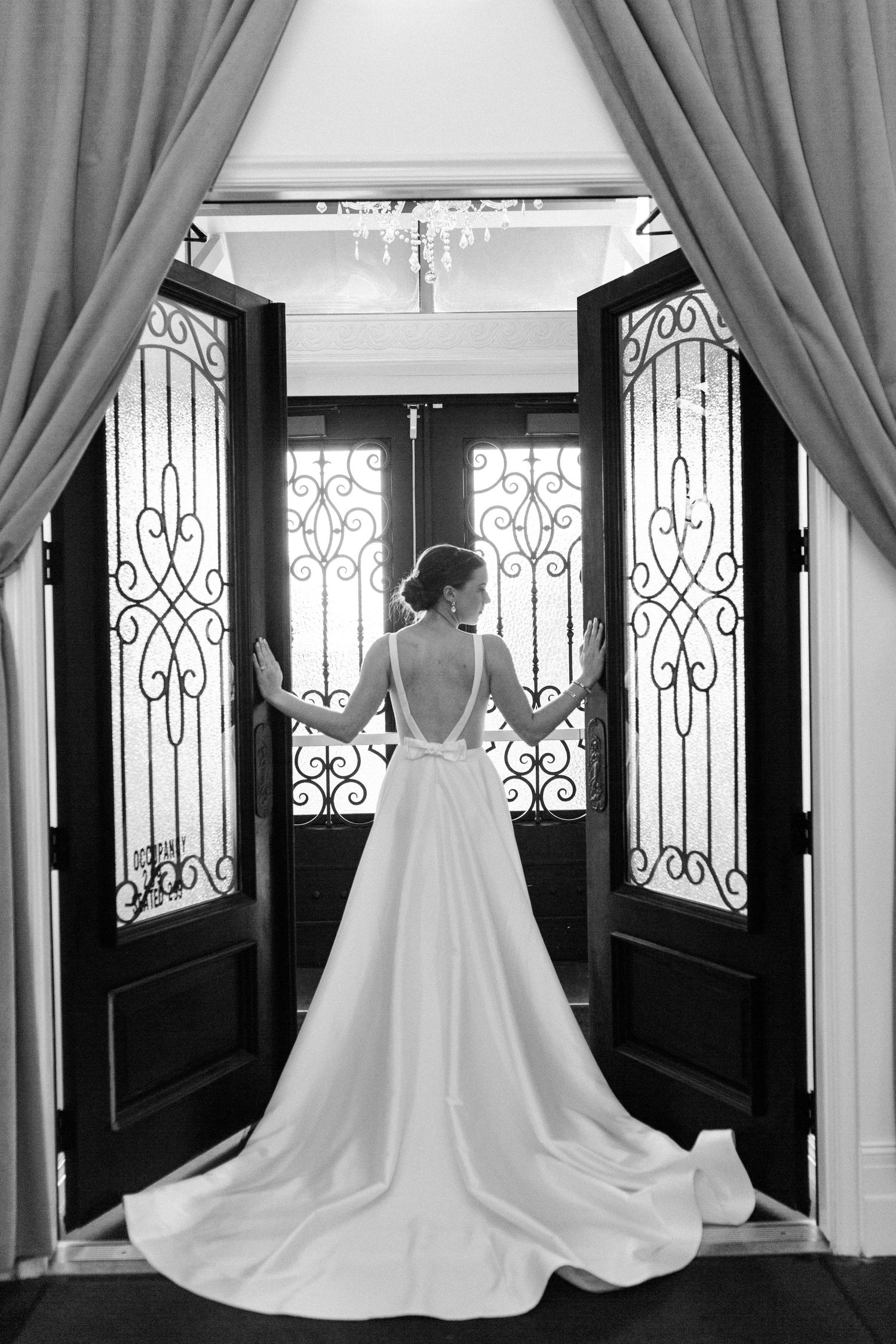 A row of three elegant white wedding gowns displayed against dark doors with ornate iron detailing and flowing curtains.