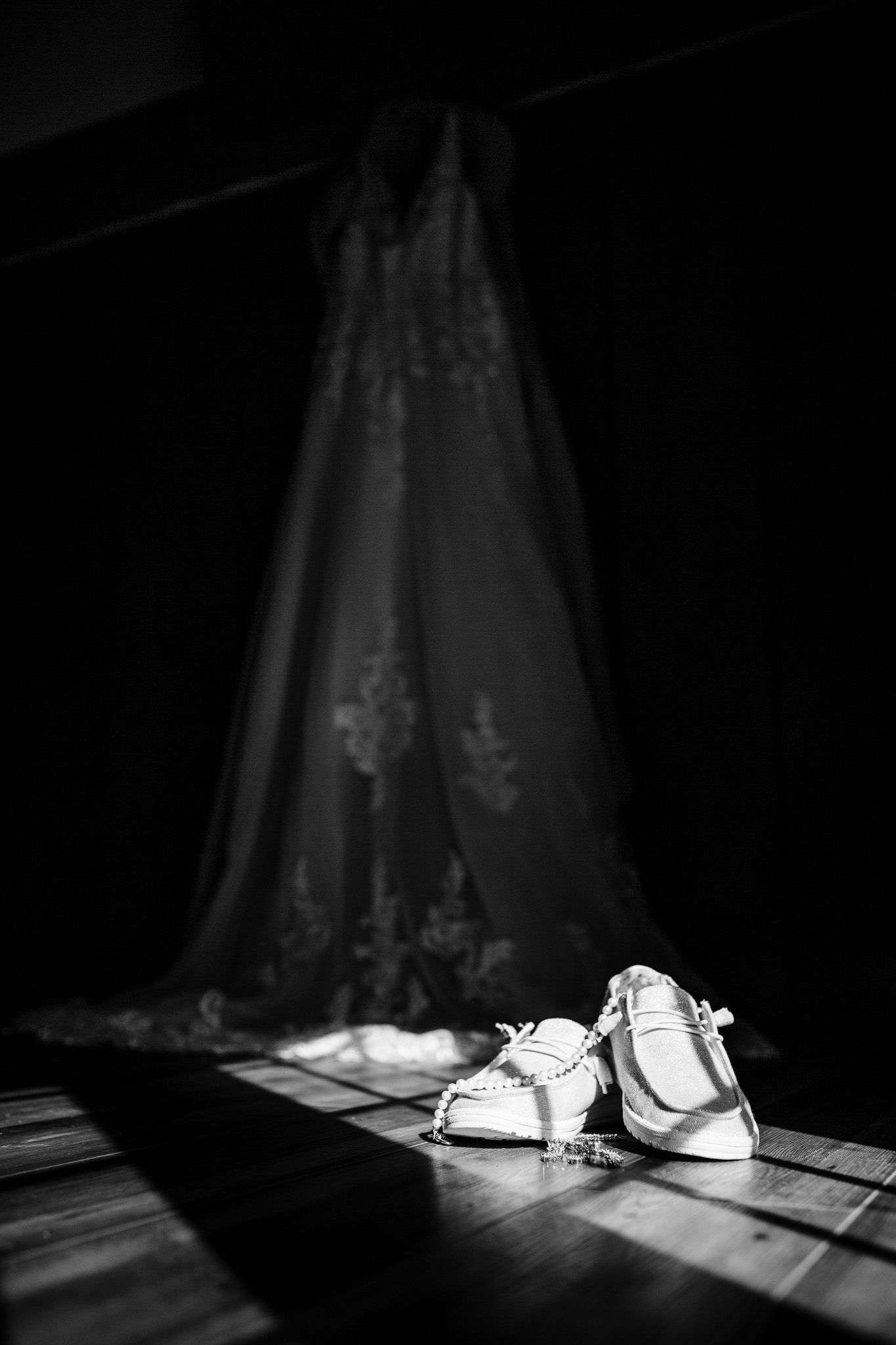 Wedding shoes and dress cast dramatic shadows in black and white lighting on a wooden floor.