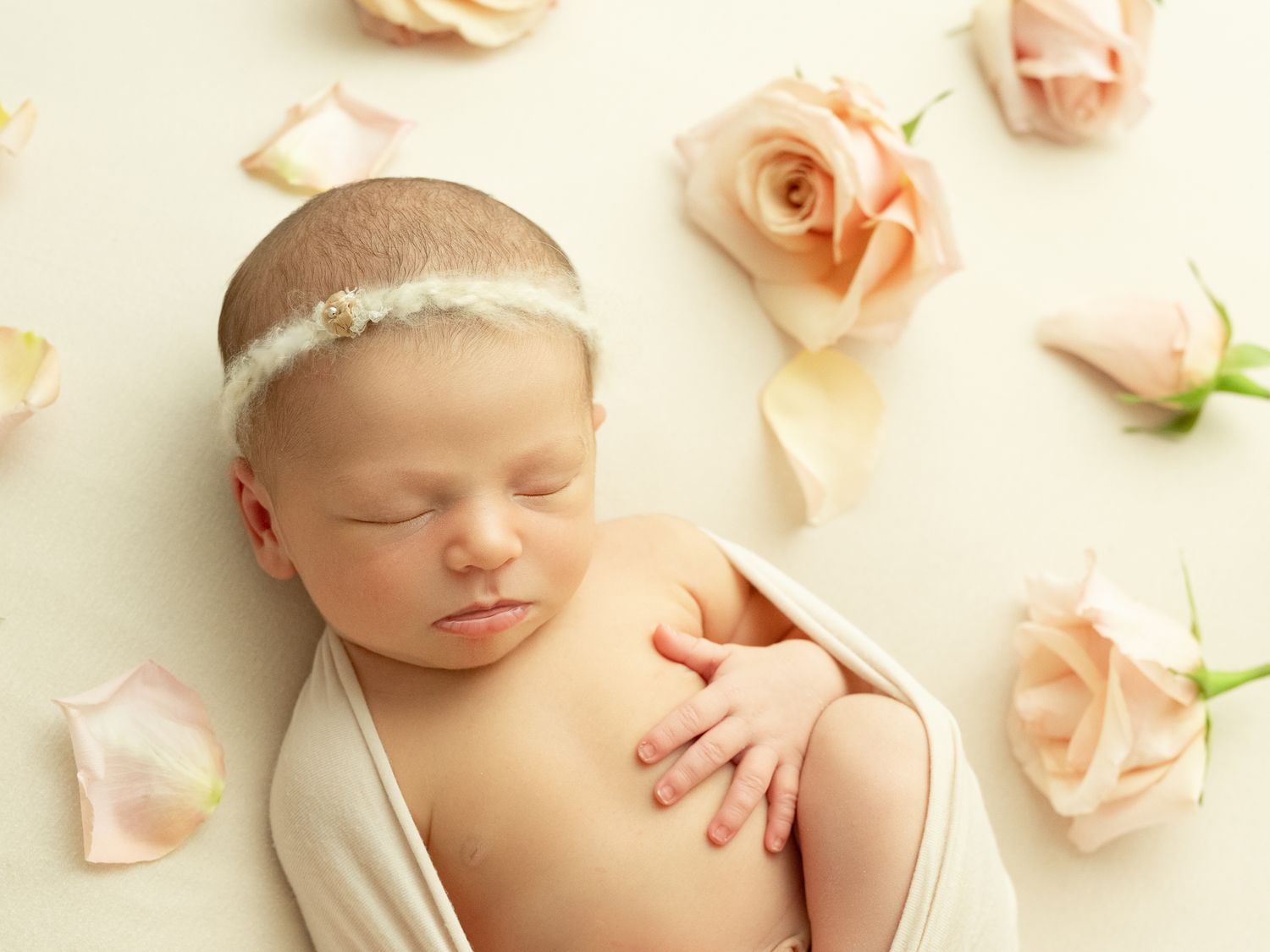 A peaceful newborn sleeping serenely surrounded by soft pink rose petals against a light neutral background.