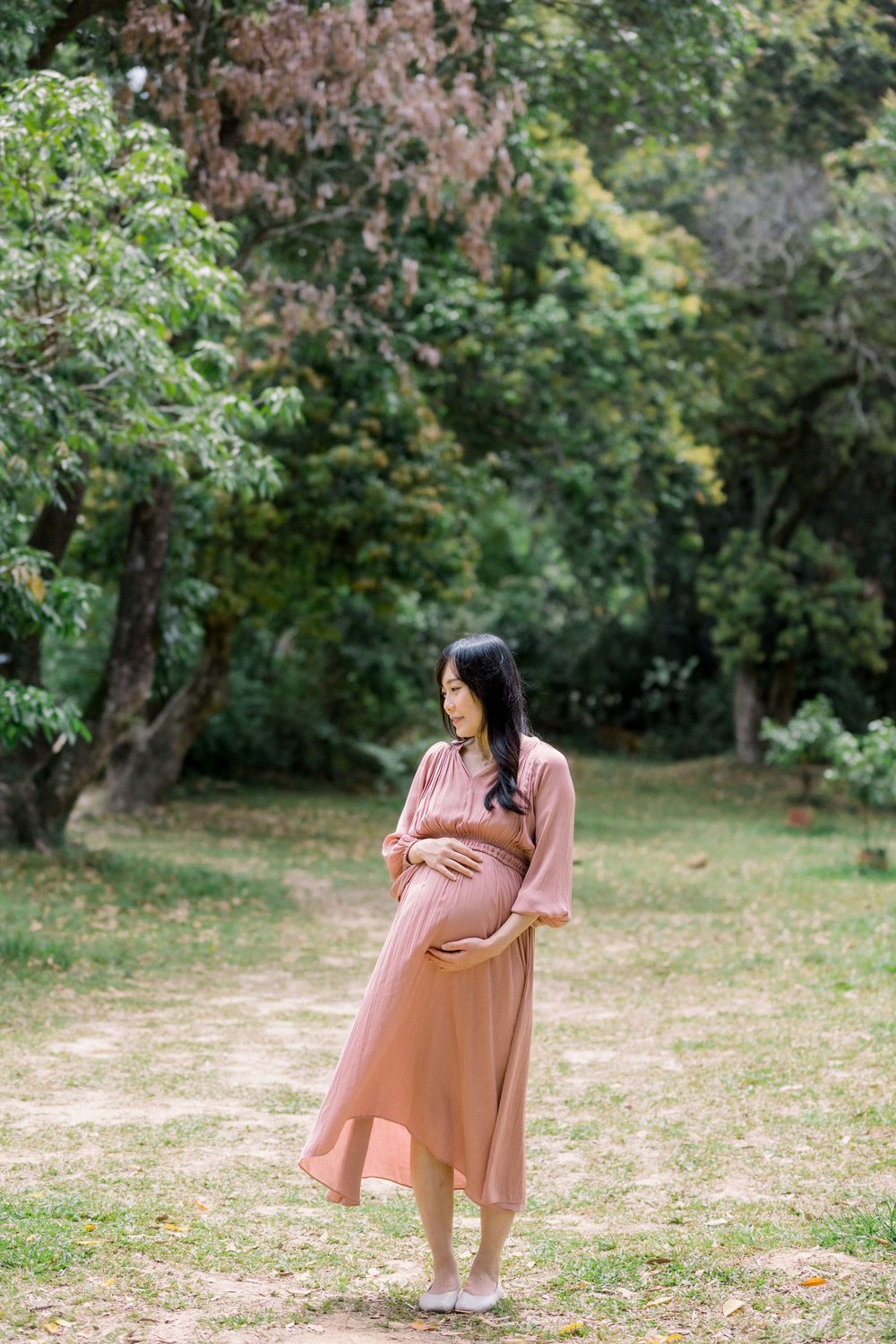 A pregnant woman in a flowing pink dress poses gracefully in a serene garden setting during autumn.