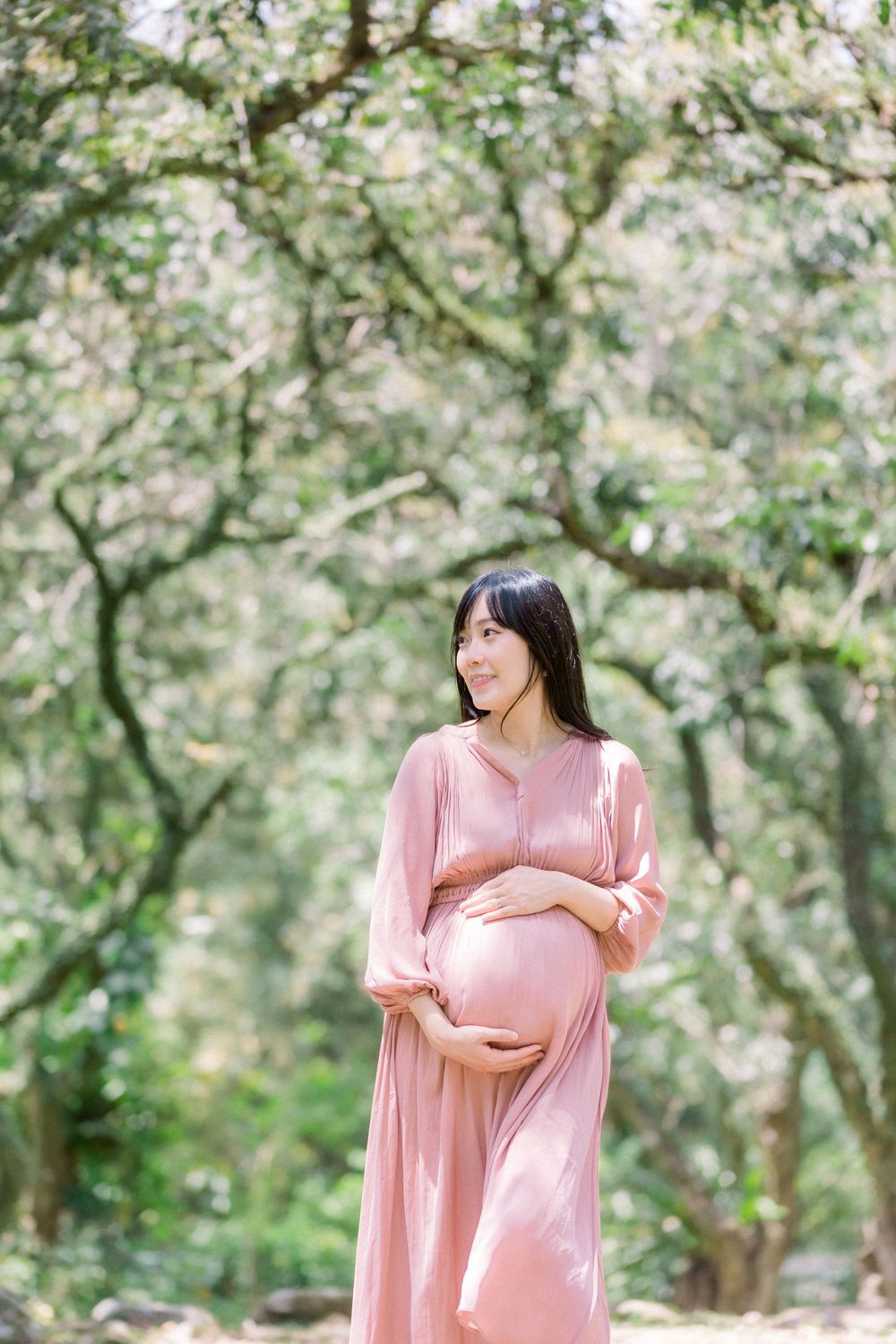 A pregnant woman in a flowing pink dress poses thoughtfully in a sunlit forest setting.