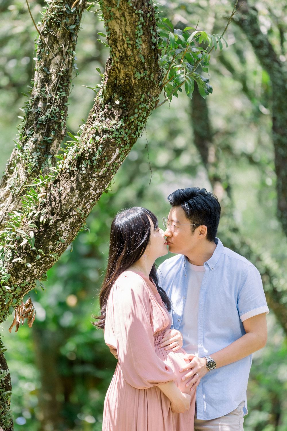 A couple shares a romantic moment in nature surrounded by trees with dappled sunlight filtering through the leaves.