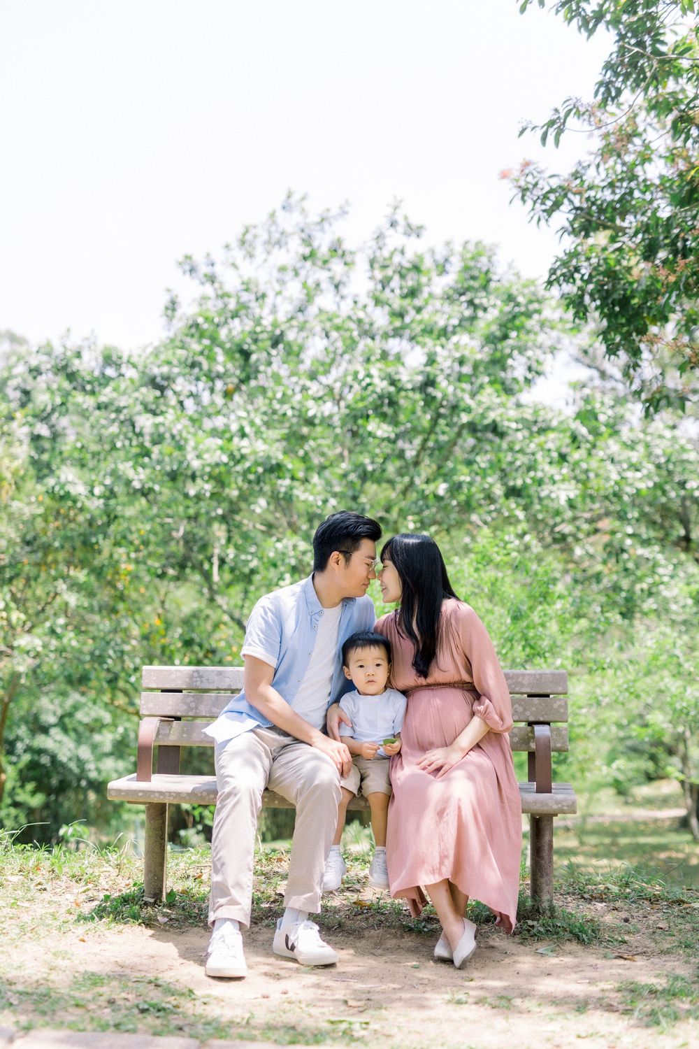 A family sits together on a wooden bench in an orchard setting surrounded by lush green trees and foliage.