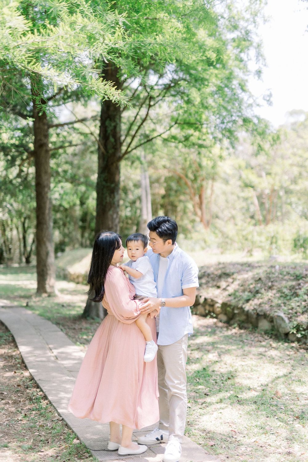 A family takes a stroll together on a tree-lined path in a sun-dappled park wearing coordinated pink and white outfits.