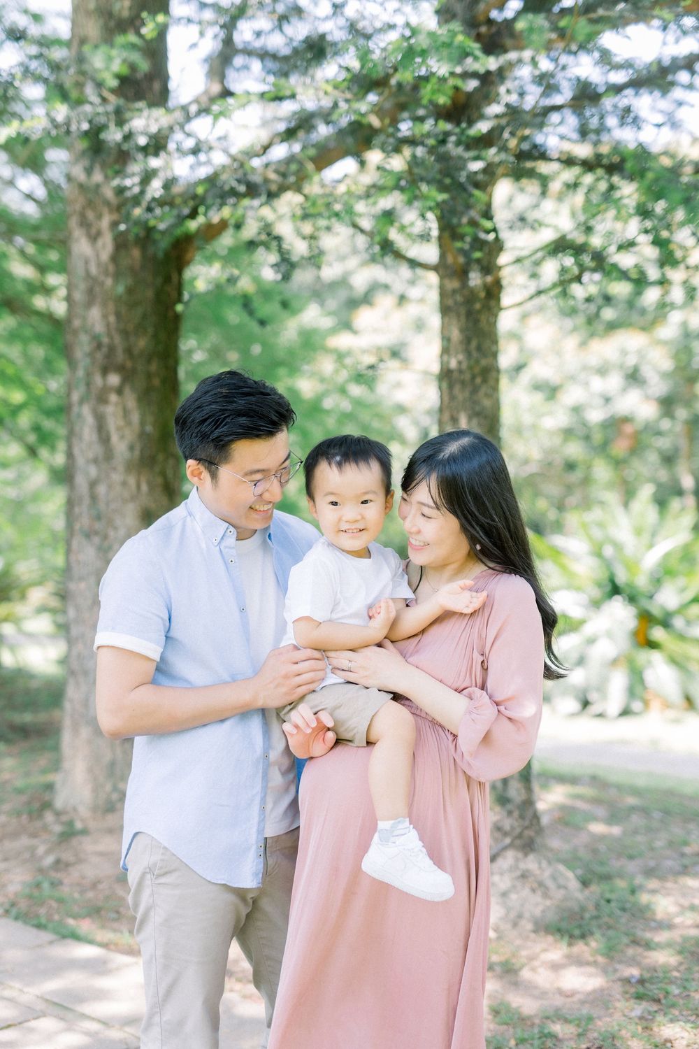 A family shares tender moments together in a sun-dappled forest setting wearing coordinated pink and white outfits.