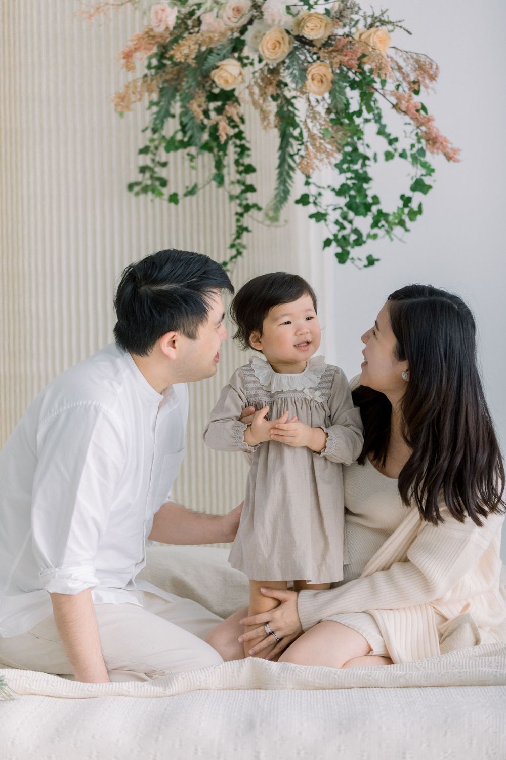 A family in white and beige outfits shares tender moments together on a couch beneath hanging greenery and florals.