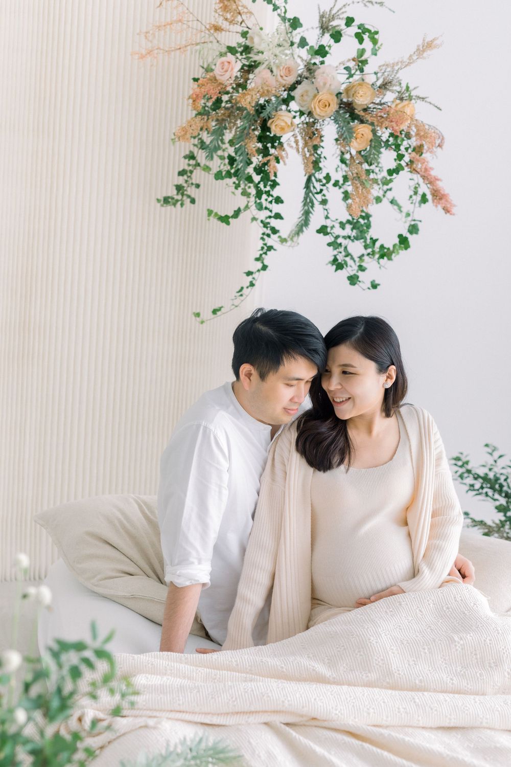 Intimate couple photo session with hanging floral arrangement and soft natural lighting in bedroom setting.