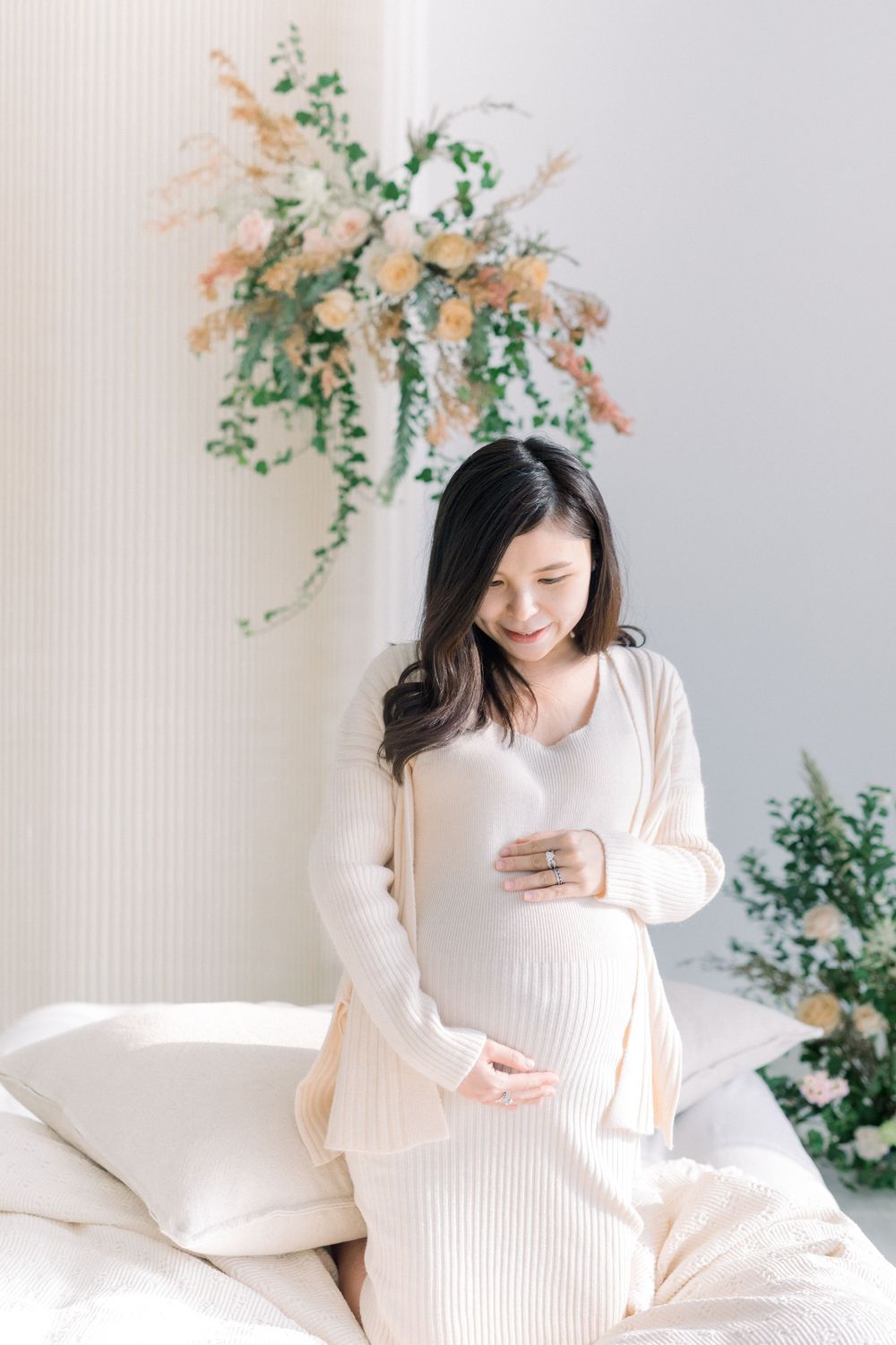 A soft and dreamy maternity photoshoot featuring a woman in white against a light background with delicate greenery.