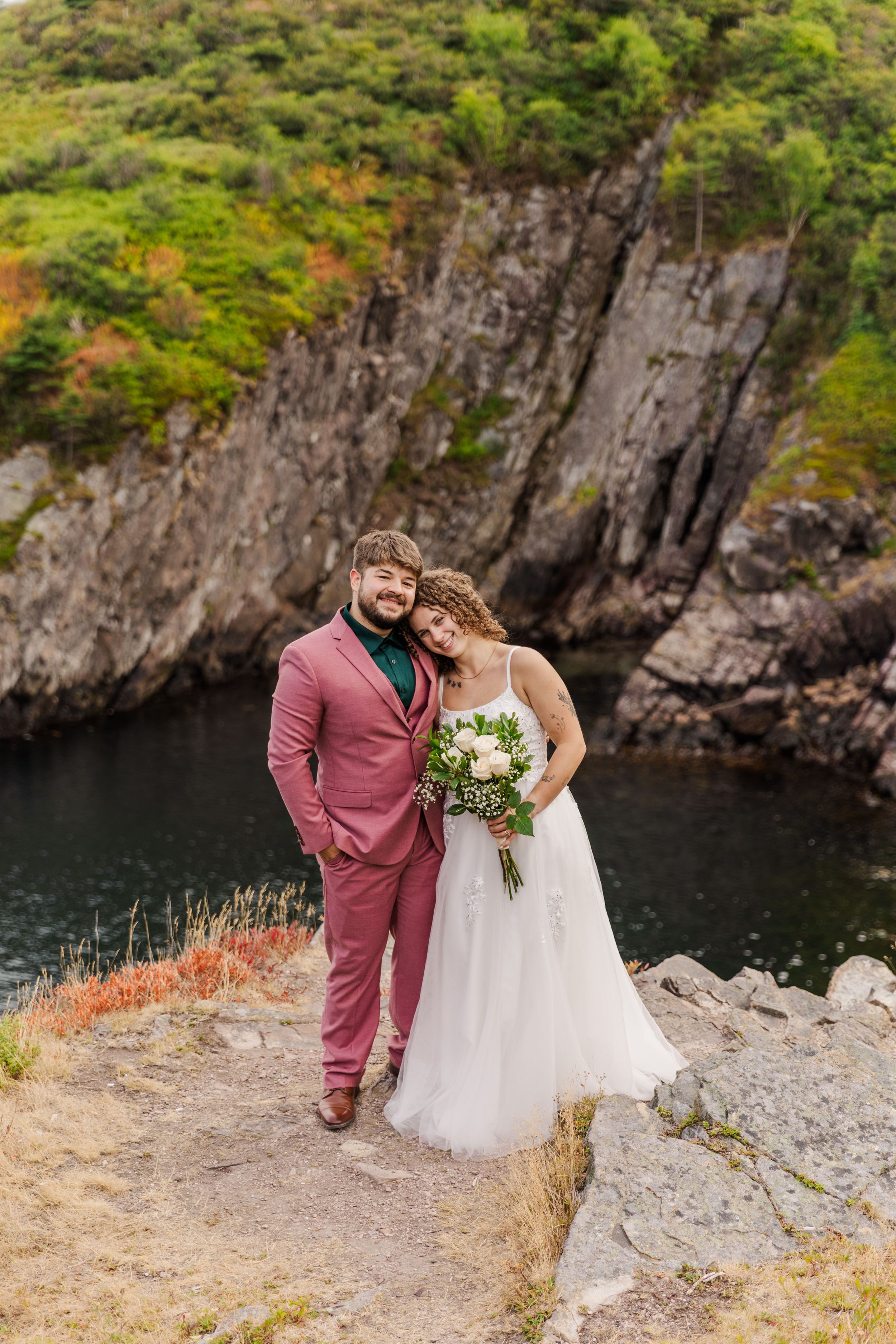 A couple in wedding attire stands at the edge of a dramatic rocky cliff overlooking water on their special day.