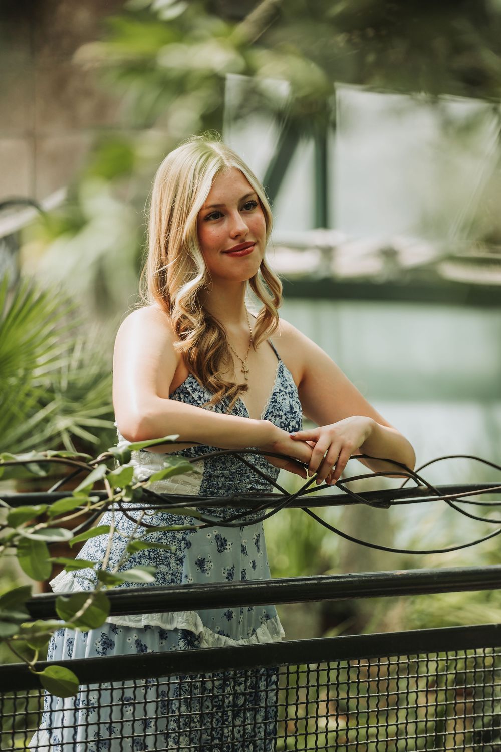 A woman in a black and white floral dress leans against a railing in an outdoor garden setting.