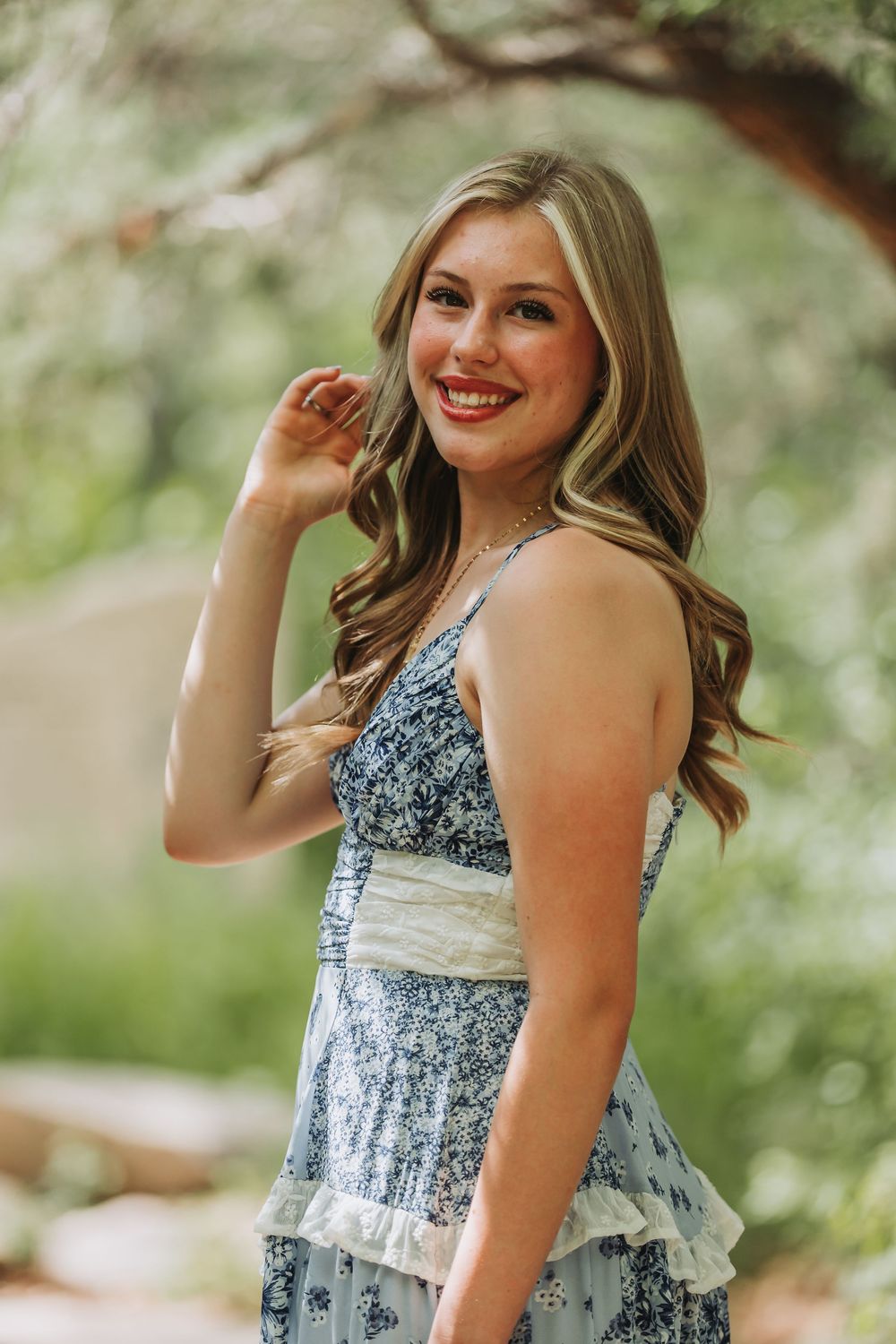 A person in a blue and white floral sundress poses outdoors in a natural garden setting on a sunny day.