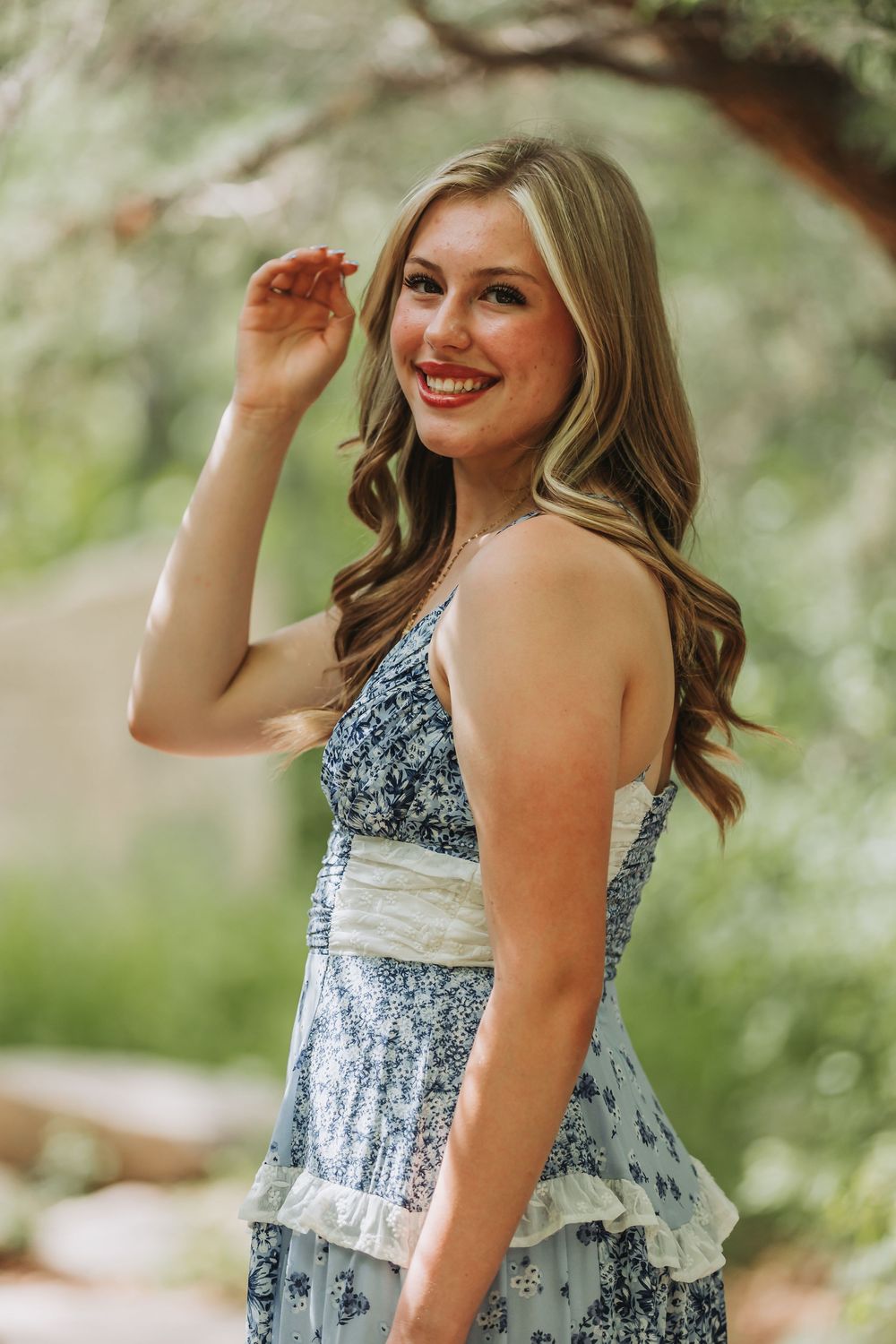 A woman in a blue and white floral sundress smiles while posing outdoors in a natural garden setting.