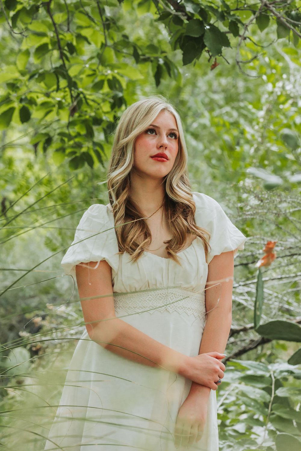 A woman in a white dress poses thoughtfully in a lush green garden setting with soft natural lighting.