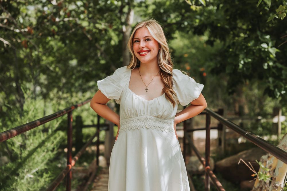 A woman in a white dress with puff sleeves poses in a sunny garden setting among lush greenery and trees.
