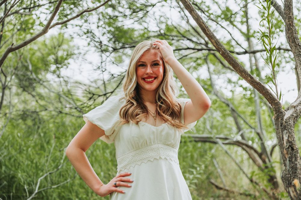 A person in a white lace wedding dress poses outdoors among trees and greenery.