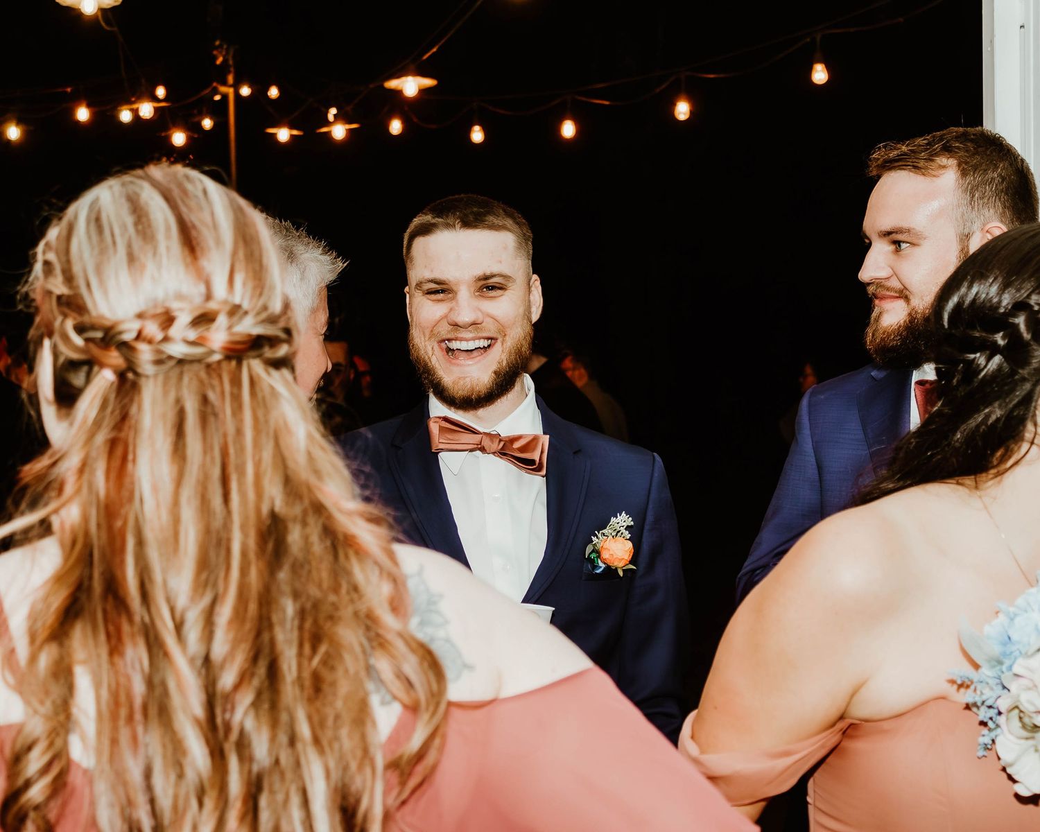 Wedding guests laugh and smile under romantic string lights during an outdoor evening reception.. Wedding party dancing with bride and groom