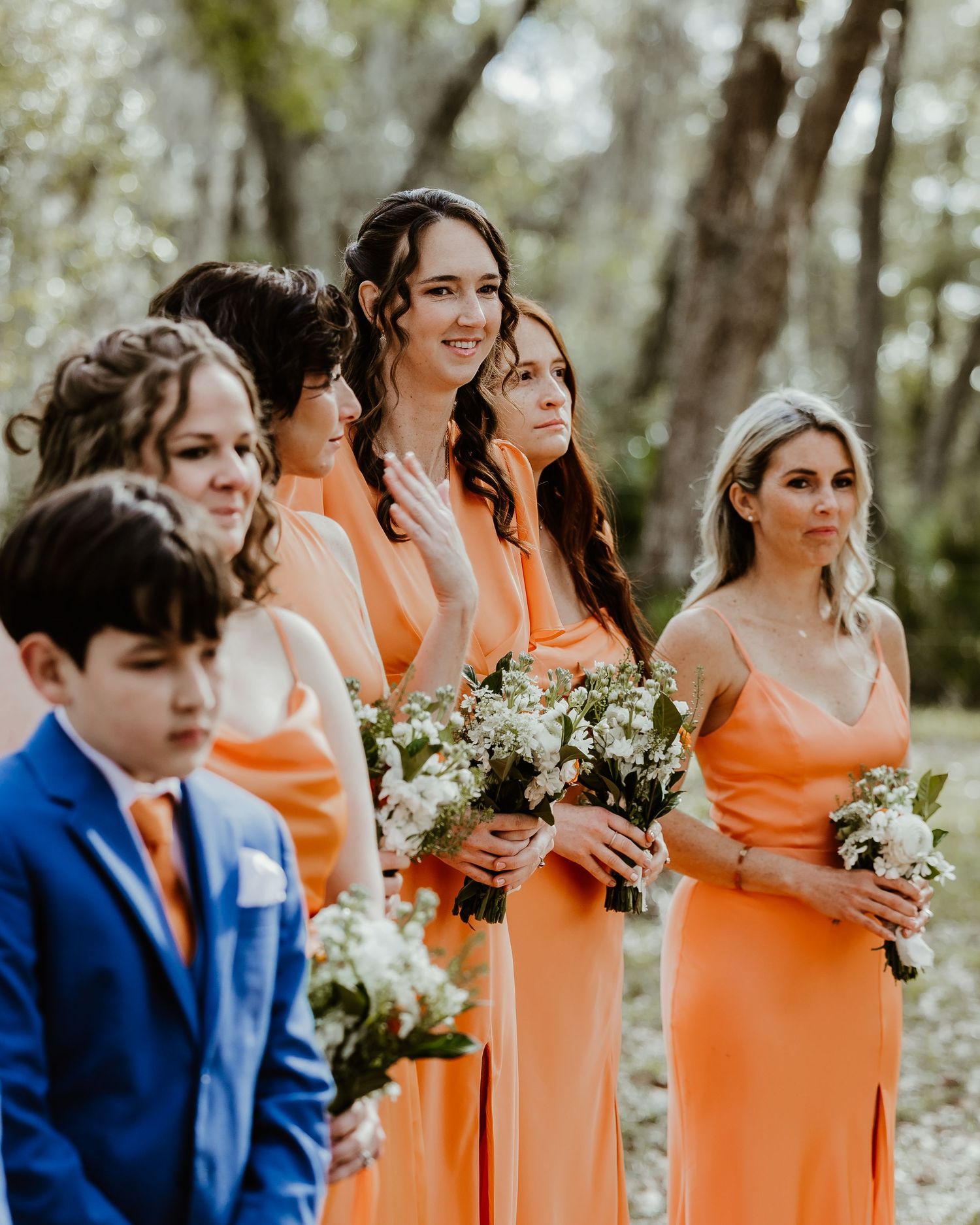 Bridesmaids wearing peach-colored dresses stand in line holding white bouquets during an outdoor wedding ceremony. Documentary wedding photo