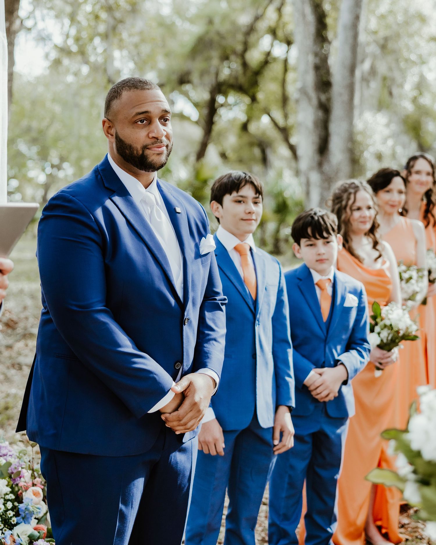 Wedding party members in navy blue suits and coral dresses stand in line during an outdoor ceremony. Groom crying at ceremony. Wedding photo