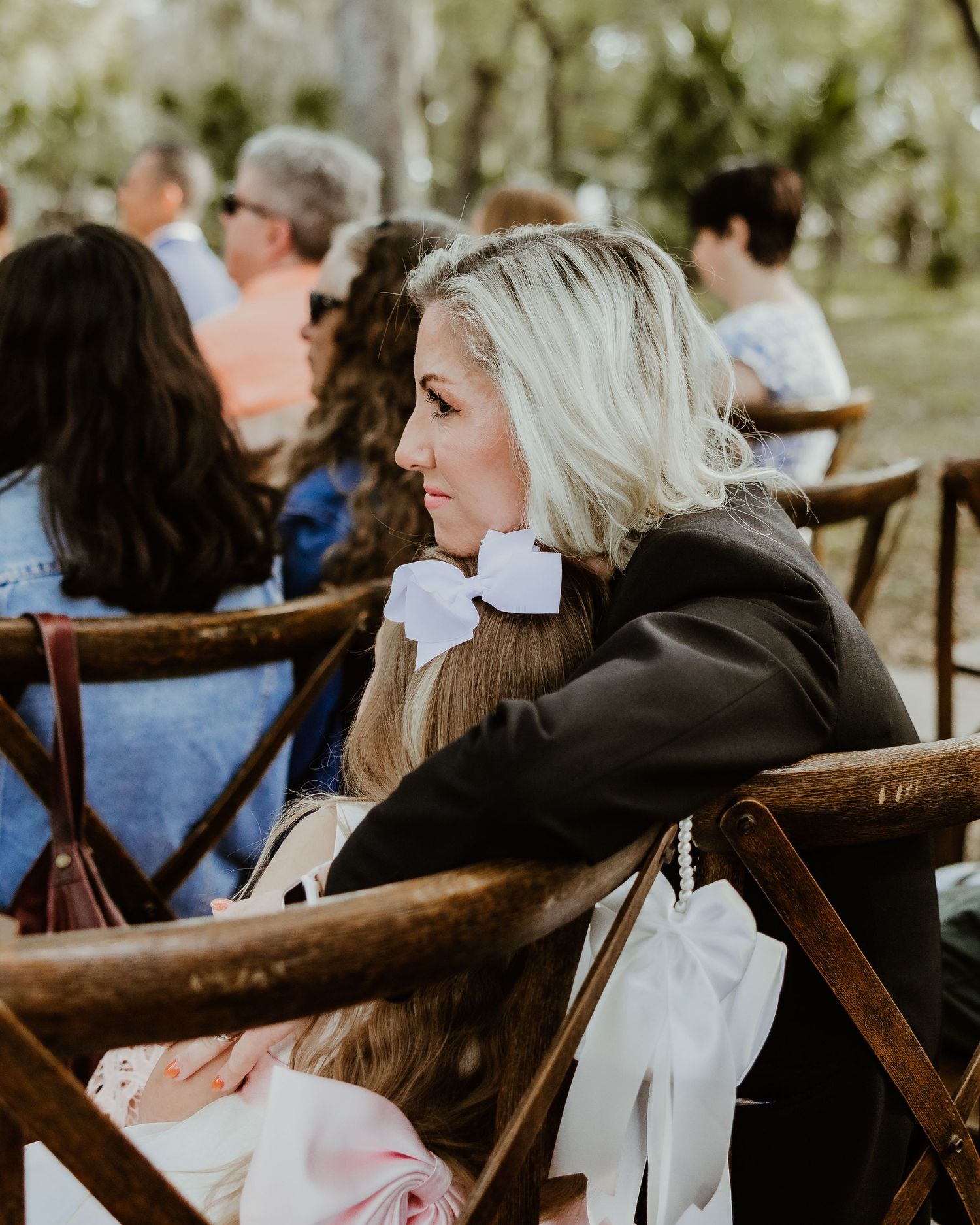 Wedding ceremony guests seated on rustic wooden chairs in an outdoor garden setting under natural sunlight. Florida barn wedding venue photo