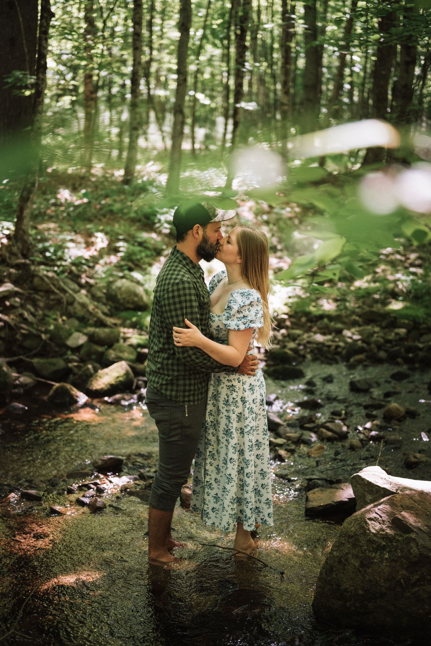 Couple sharing a kiss while standing barefoot in a forest creek surrounded by lush greenery.