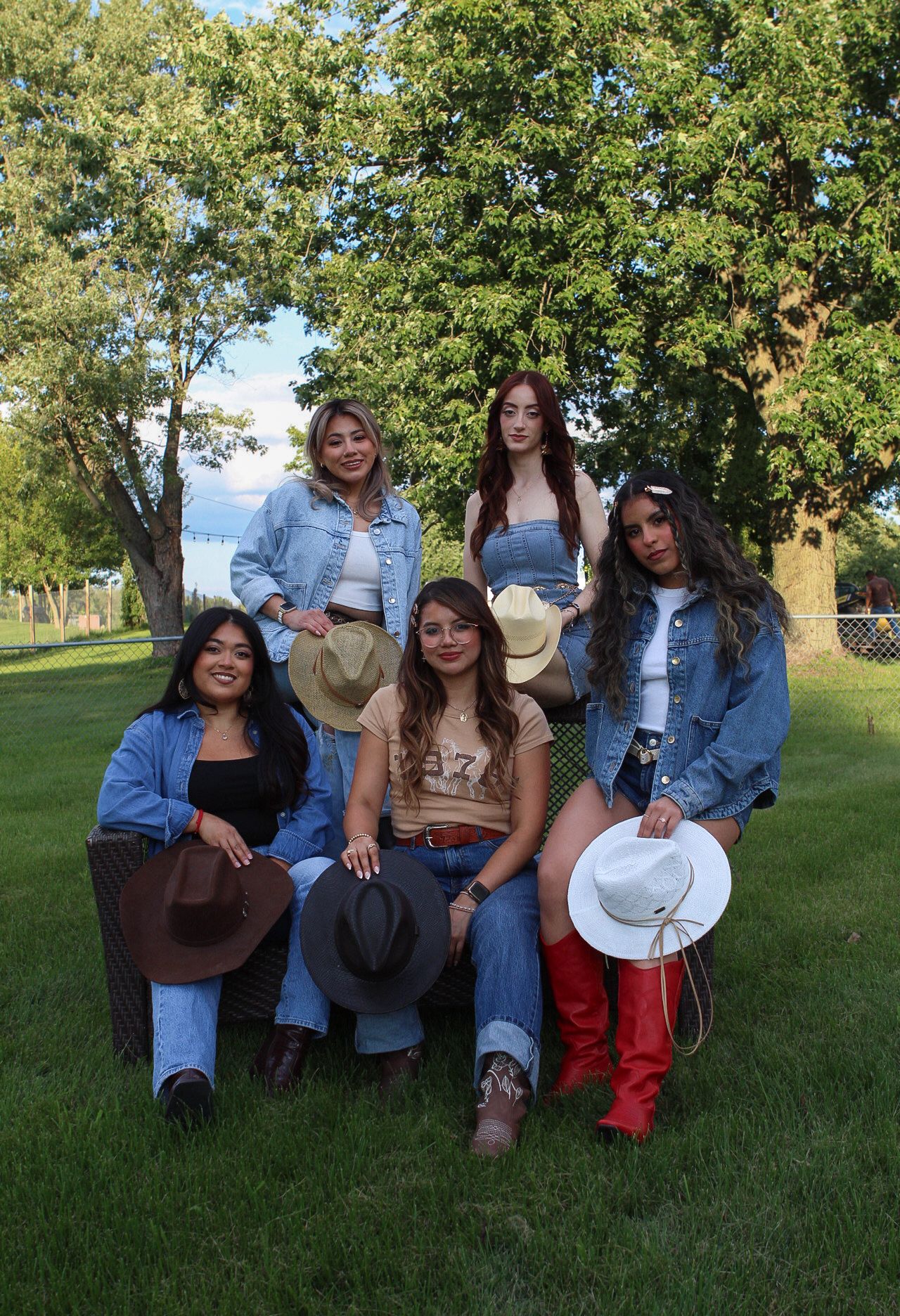 A group in western-inspired outfits with denim and cowboy hats pose together on grass during golden hour.