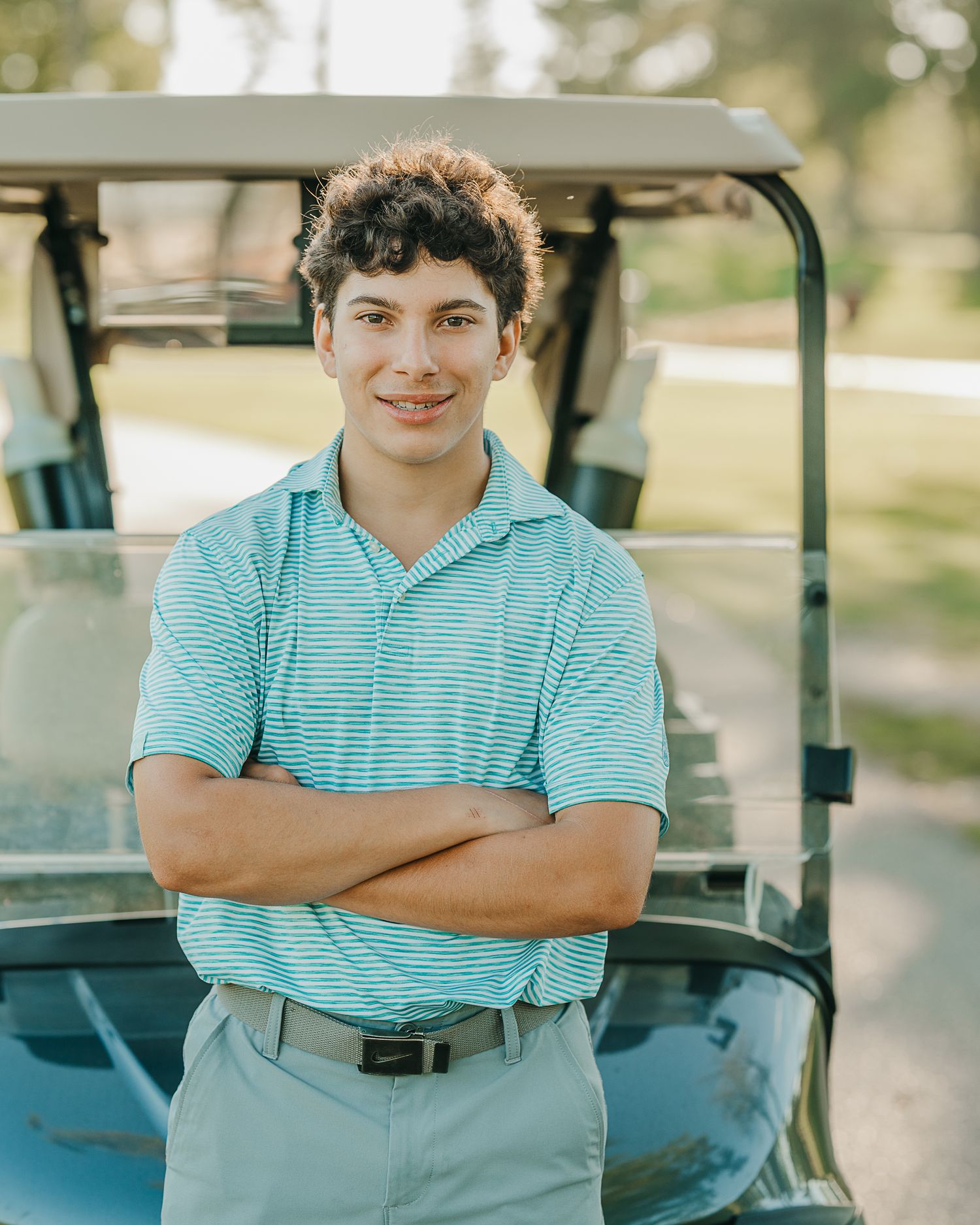 Senior golfer in a striped turquoise polo shirt stands with crossed arms by a golf cart on a sunny day in Southern New Hampshire