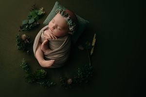 Artistic portrait of a sleeping newborn in tan wrap against dark background with greenery accents.