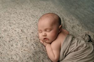 Peaceful newborn baby sleeping on textured beige backdrop wrapped in soft tan fabric.