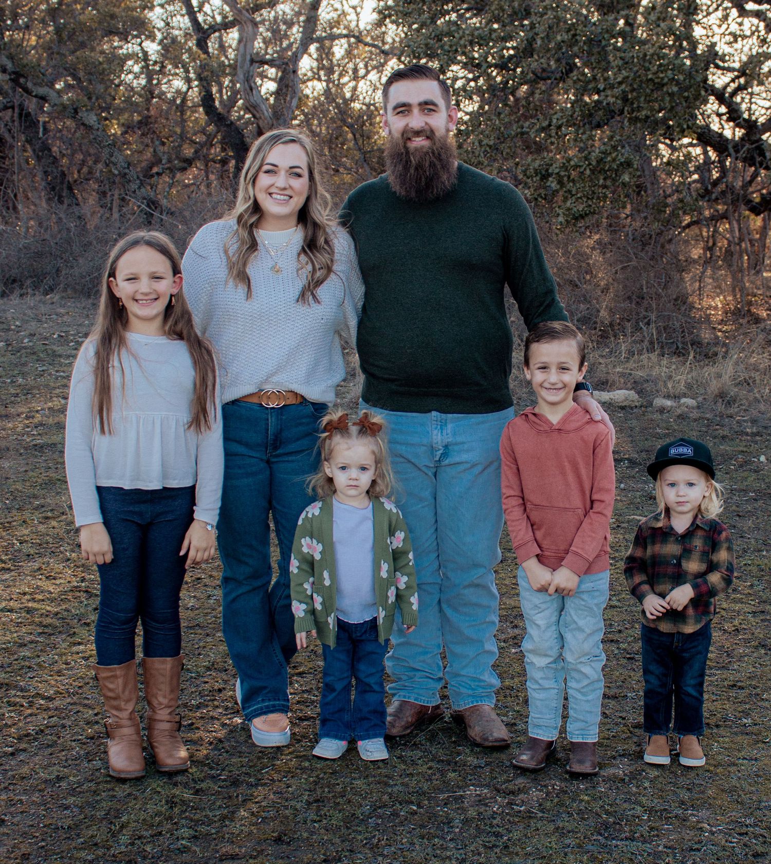 Family poses together outdoors at sunset in coordinated casual outfits against wooded backdrop.