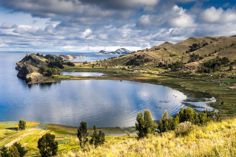 Panoramic view of Lake Titicaca with rolling hills, blue skies, and traditional Andean farmland along its shores.
