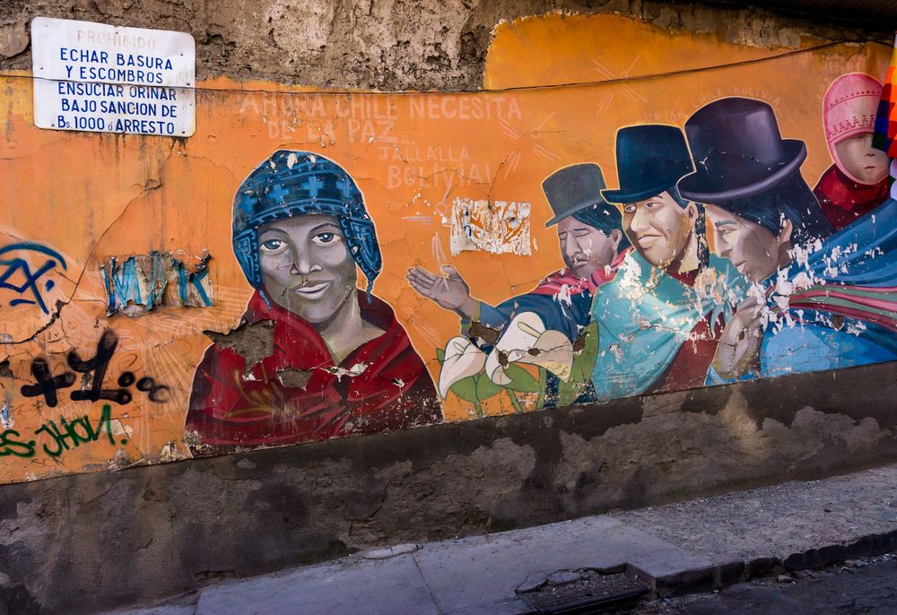 Colorful street art murals adorn an orange wall along a sidewalk in an urban setting.