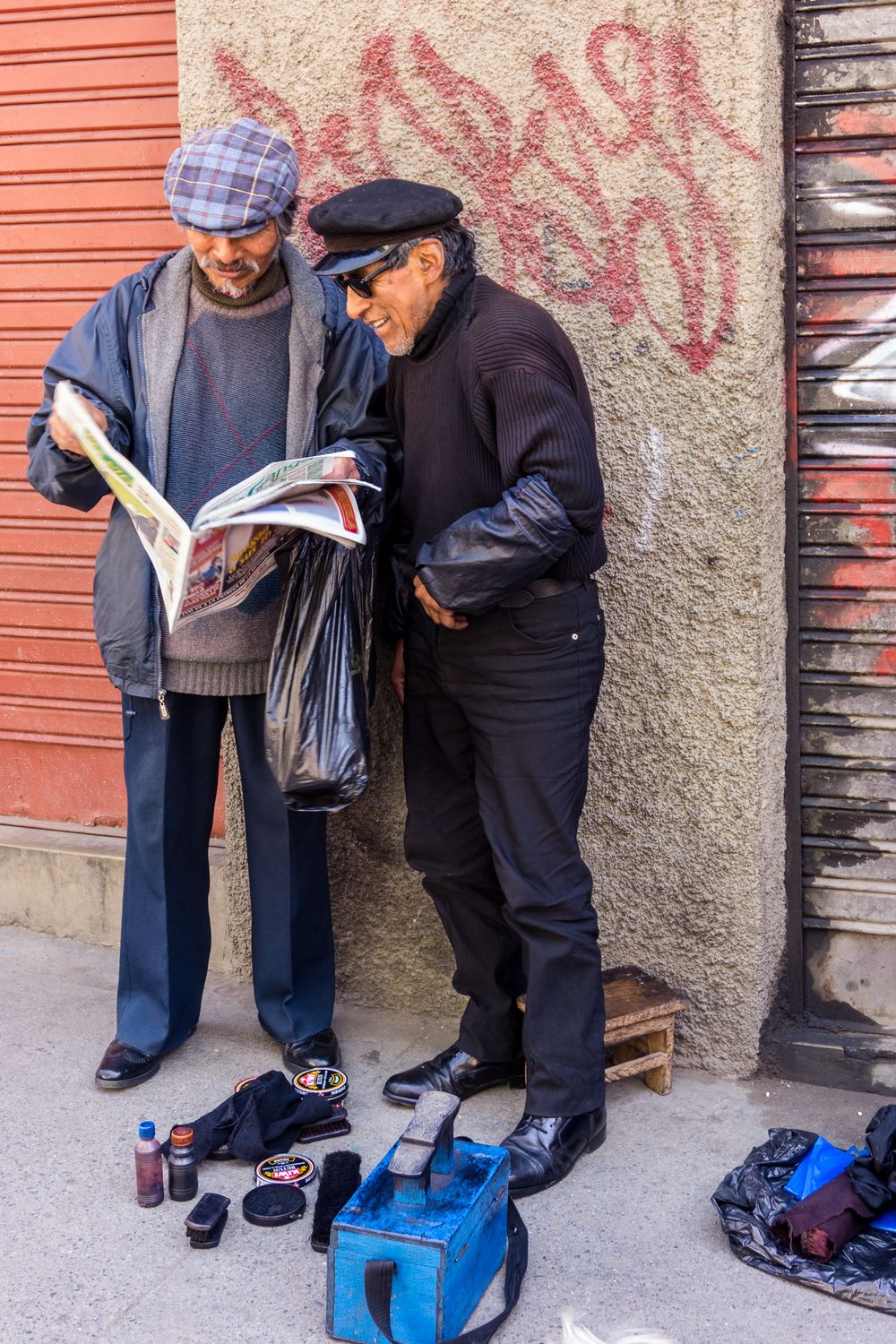 Two men stand closely together reading documents against an orange wall with graffiti.