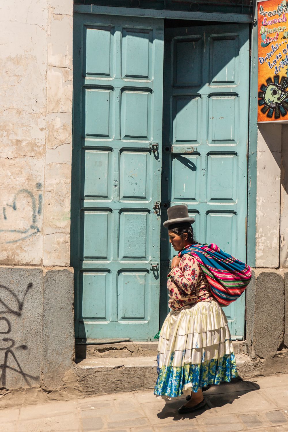 Indigenous women in traditional dress walk past turquoise doors on a stone building in Peru.