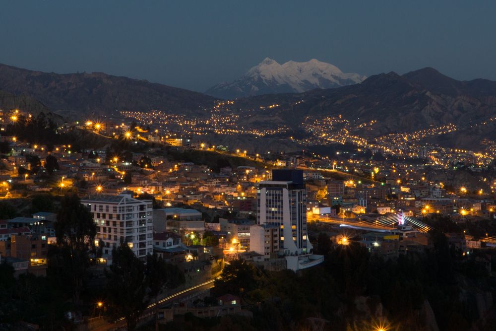 Panoramic night view of a city with glowing lights nestled in mountainous terrain and snowy peaks in the distance.