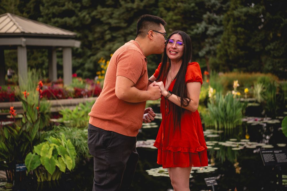 A couple celebrates their engagement in a garden setting with a pond and lush greenery in the background.