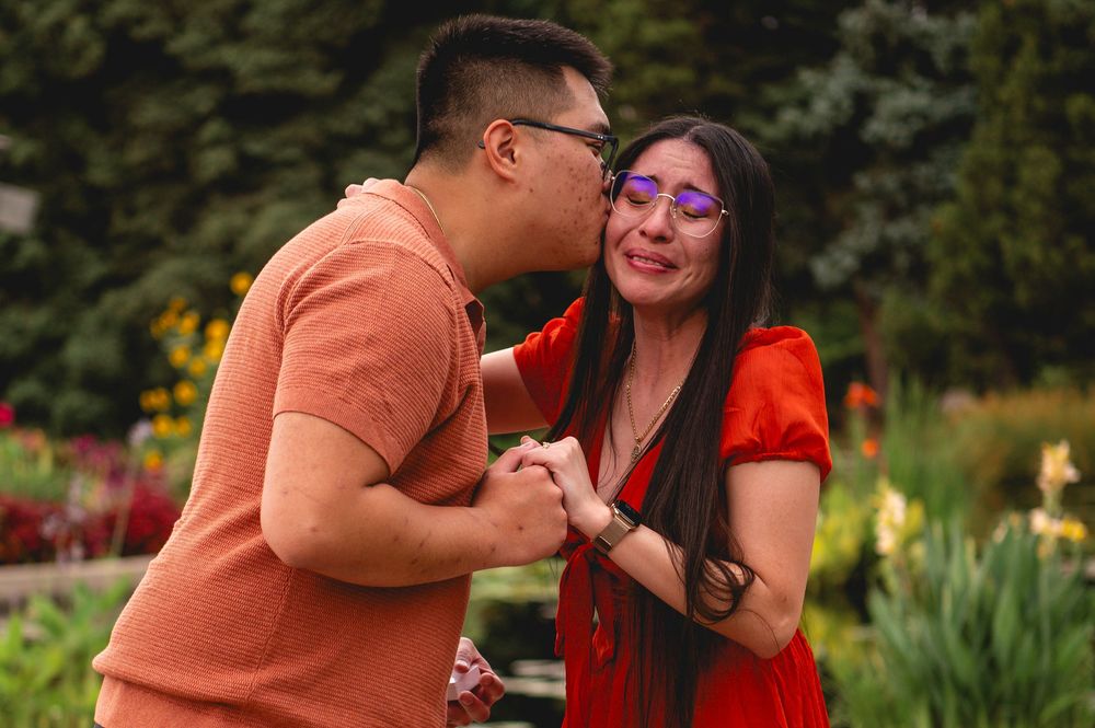 A couple in orange and red outfits embrace outdoors in front of trees and greenery during a portrait session.