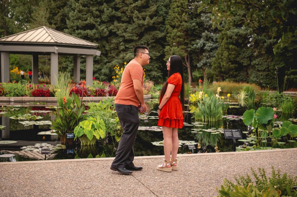 Two people share a romantic moment near a gazebo in a lush garden with red flowers and lily ponds.